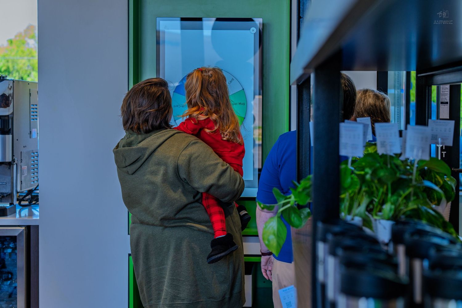 Woman holding child views a large interactive digital display screen inside a modern green retail store.