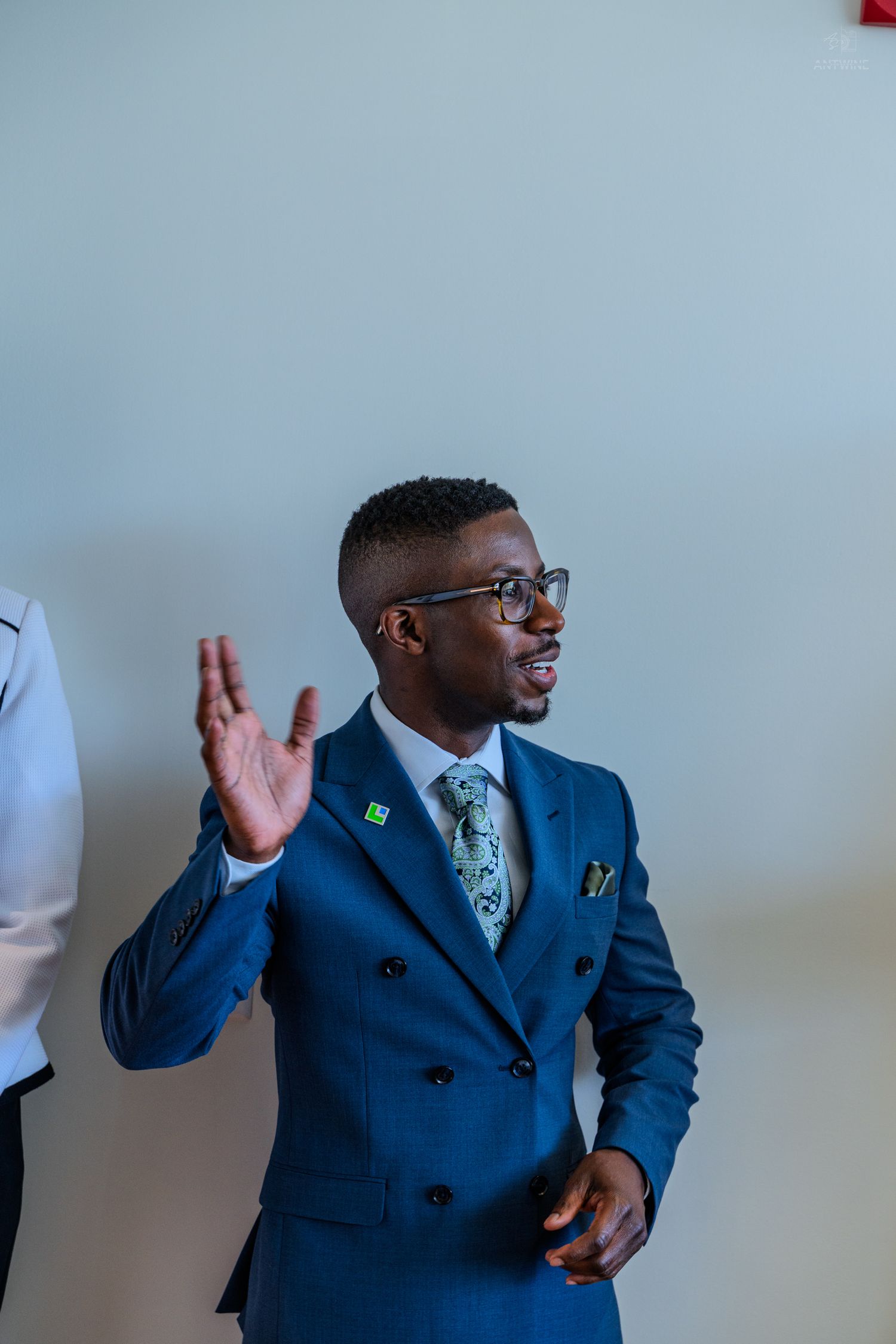 Man in blue double-breasted suit and glasses waving hand indoors.