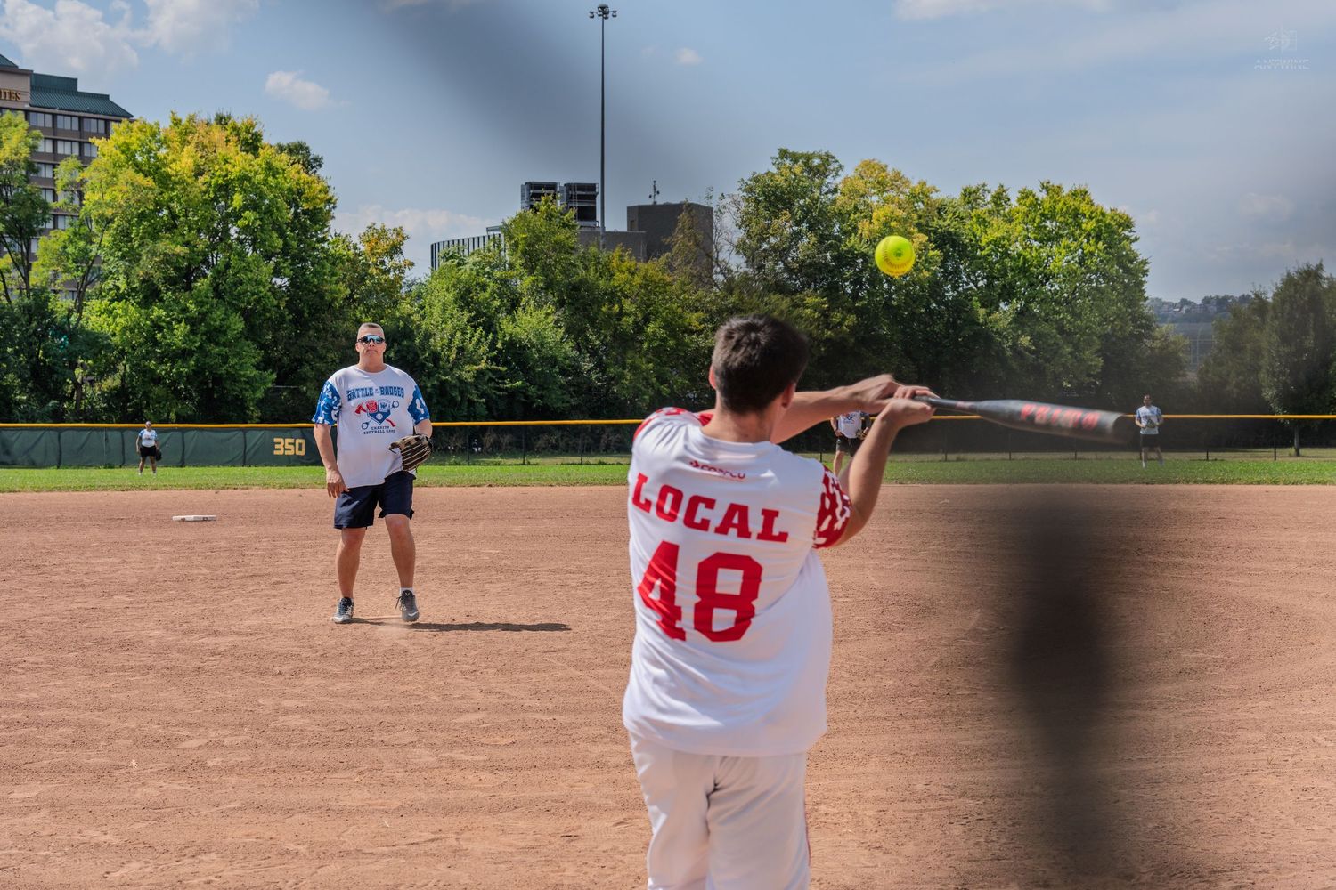 Player in Local 48 jersey swings bat at softball on dirt field during outdoor recreational game.