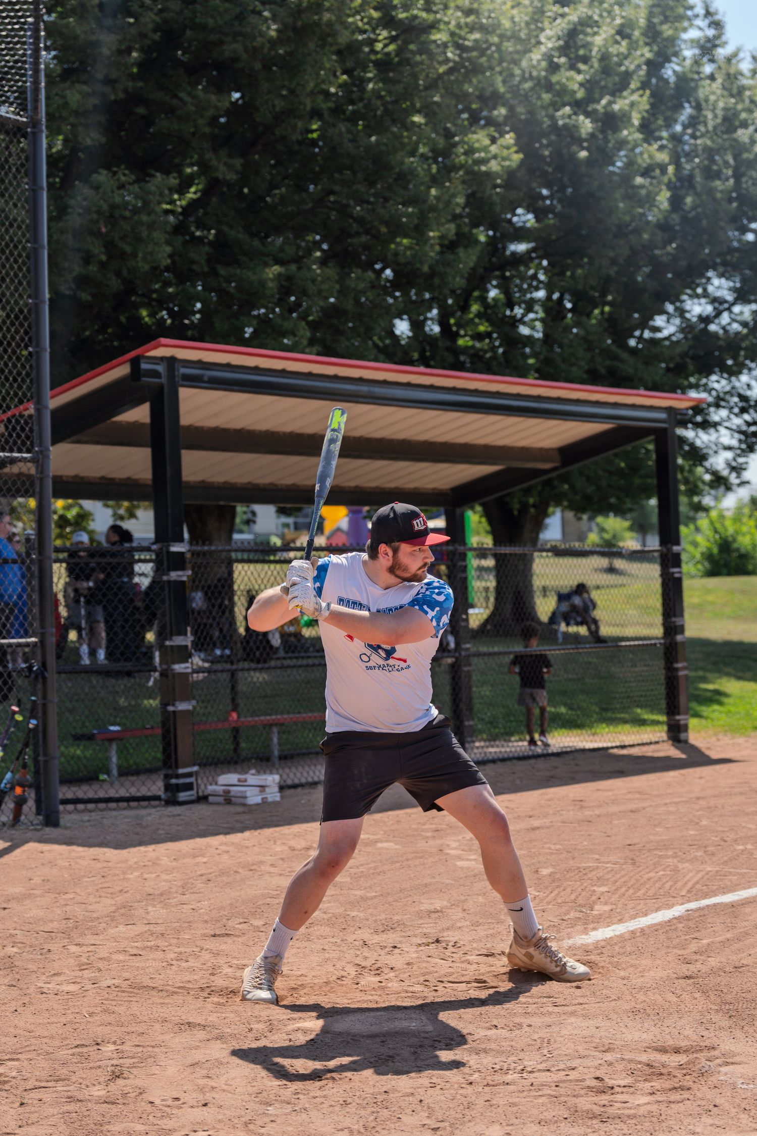 A batter stands ready at home plate during an outdoor softball game on a sunny day near a dugout.