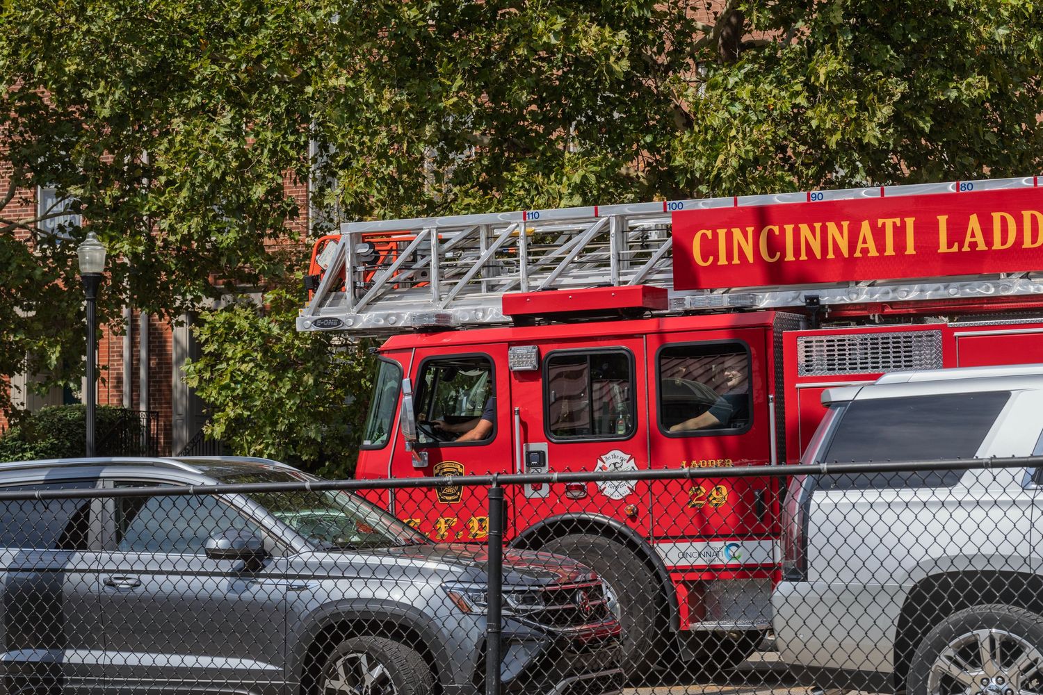 A Cincinnati Ladder fire truck parked near a chain-link fence surrounded by trees.