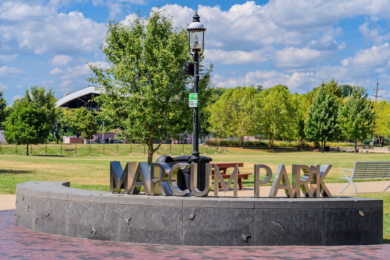 Marcus Park sign with lamp post surrounded by lush green trees and brick pathway on a sunny day.