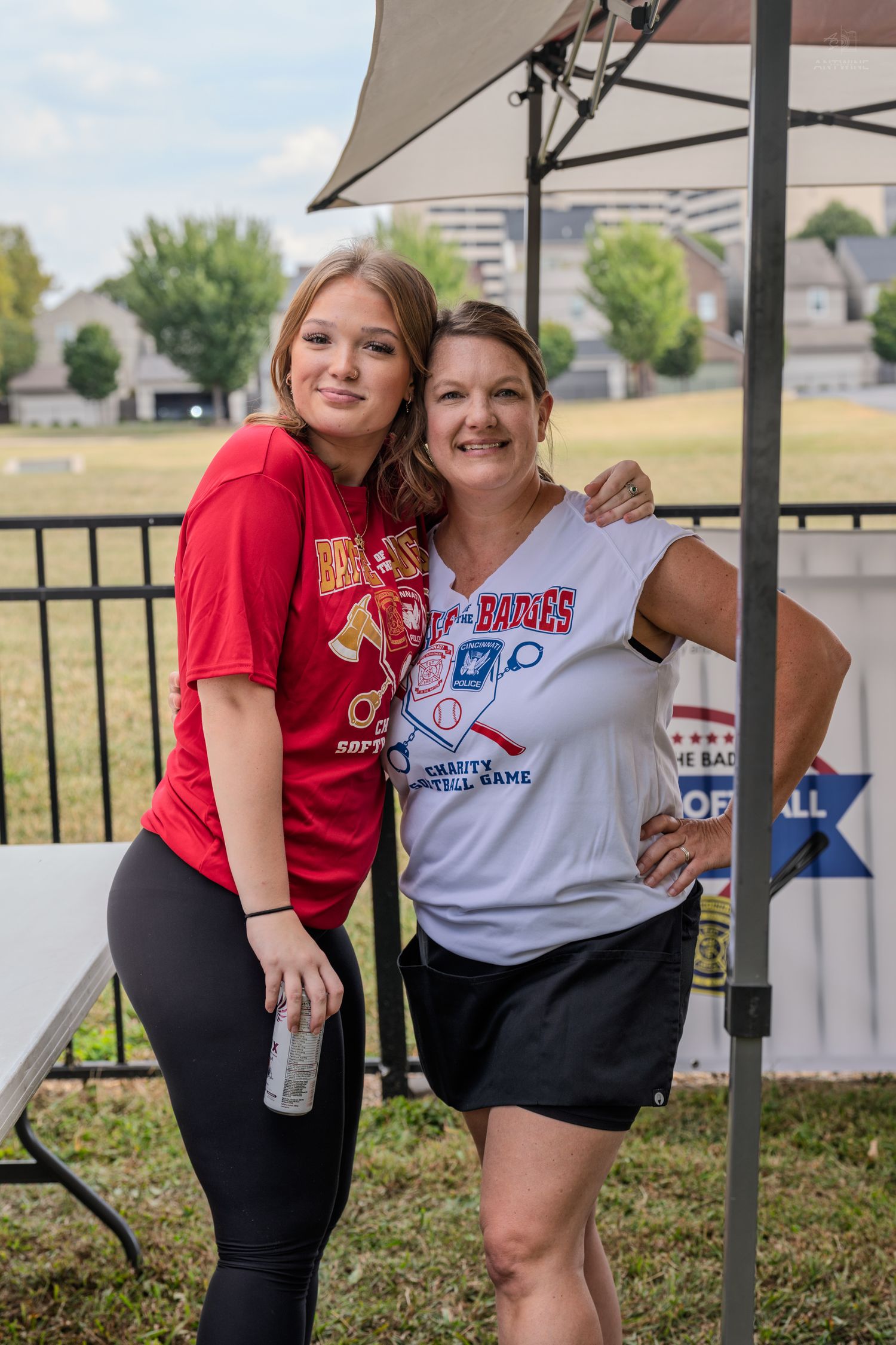 Two women smile and pose together under a tent at a charity softball game event on a sunny day.