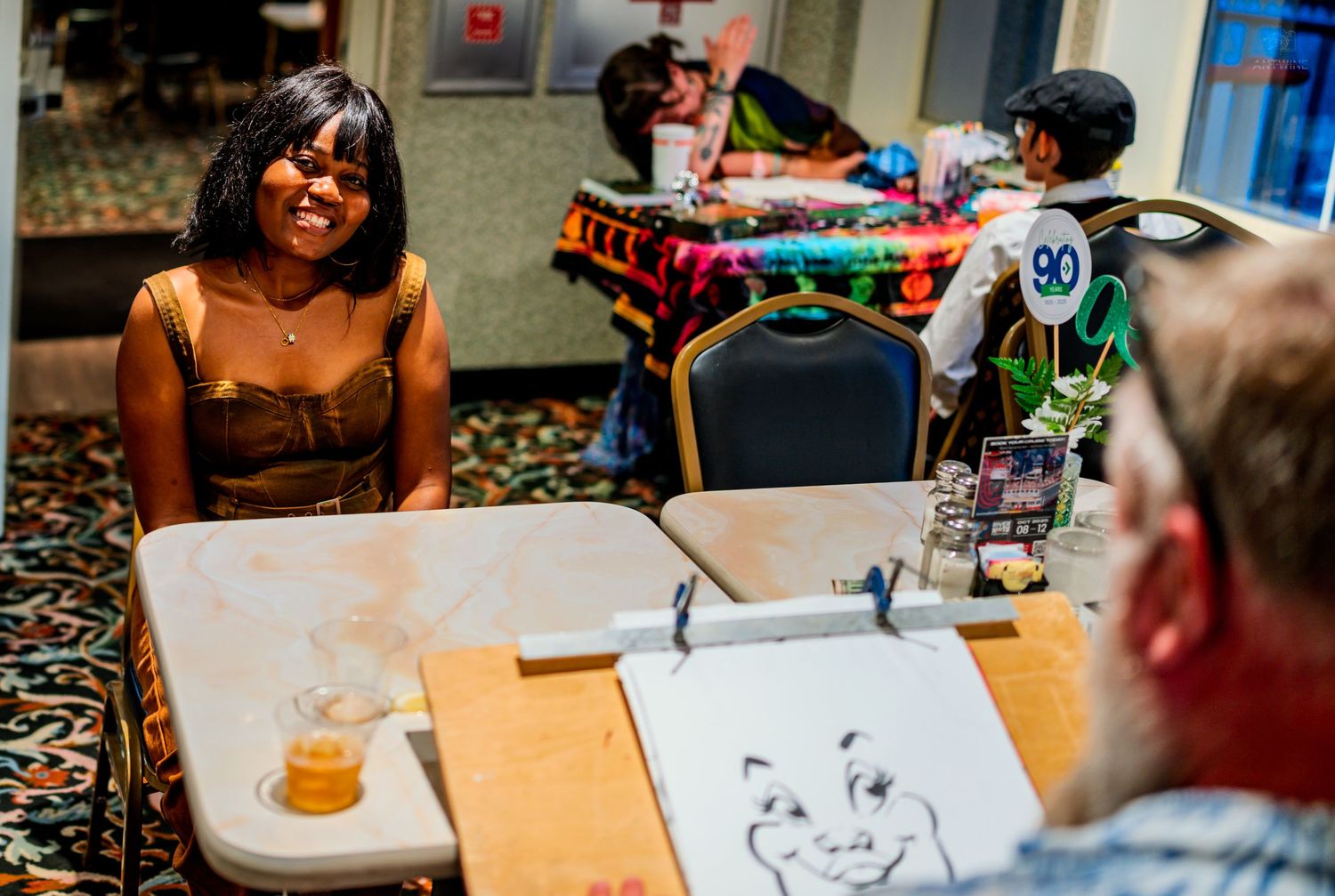 People socializing at a casual indoor event with caricature art, drinks on tables, and colorful decorations in background.