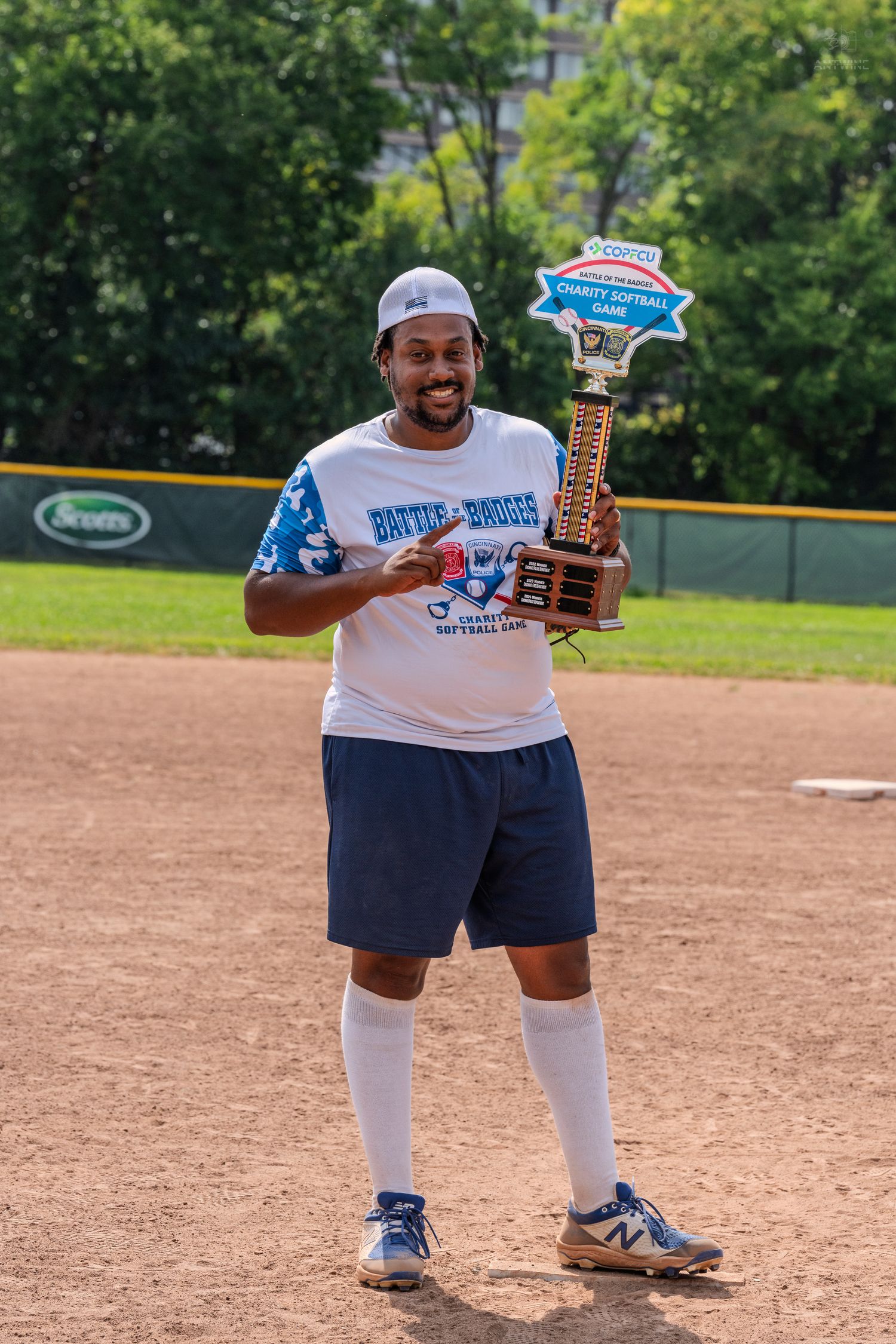 A smiling man in a blue softball jersey proudly holds a charity softball game championship trophy on a field.