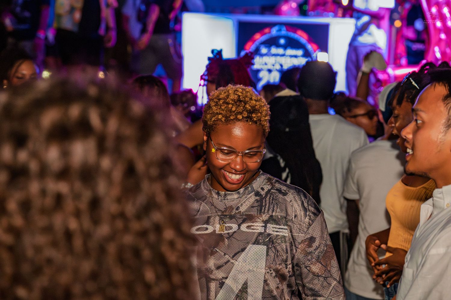 Smiling woman in Adidas jacket enjoying Juneteenth Block Party event under colorful venue lights.