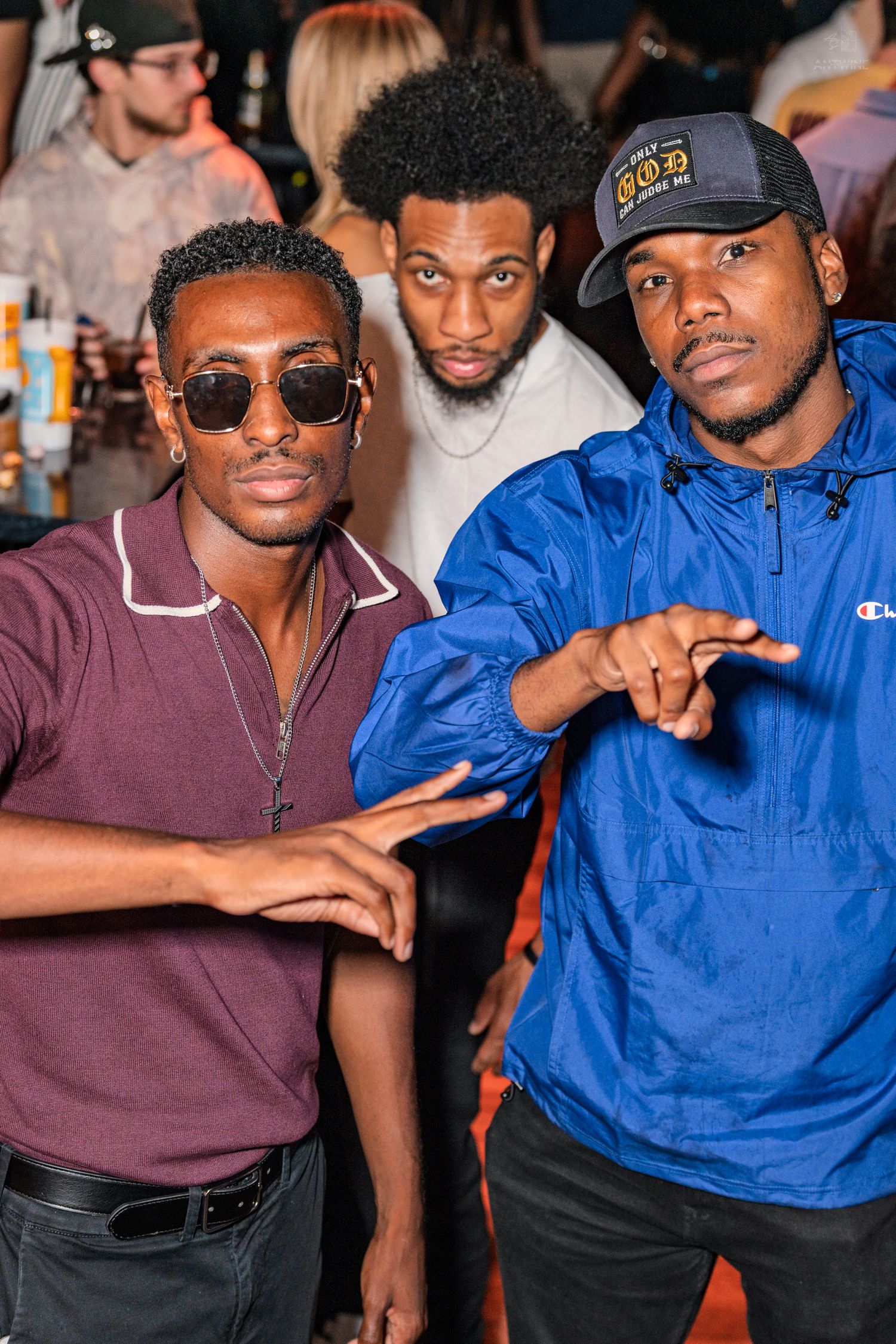 Three young men posing together at a social gathering, two making hand gestures toward the camera.