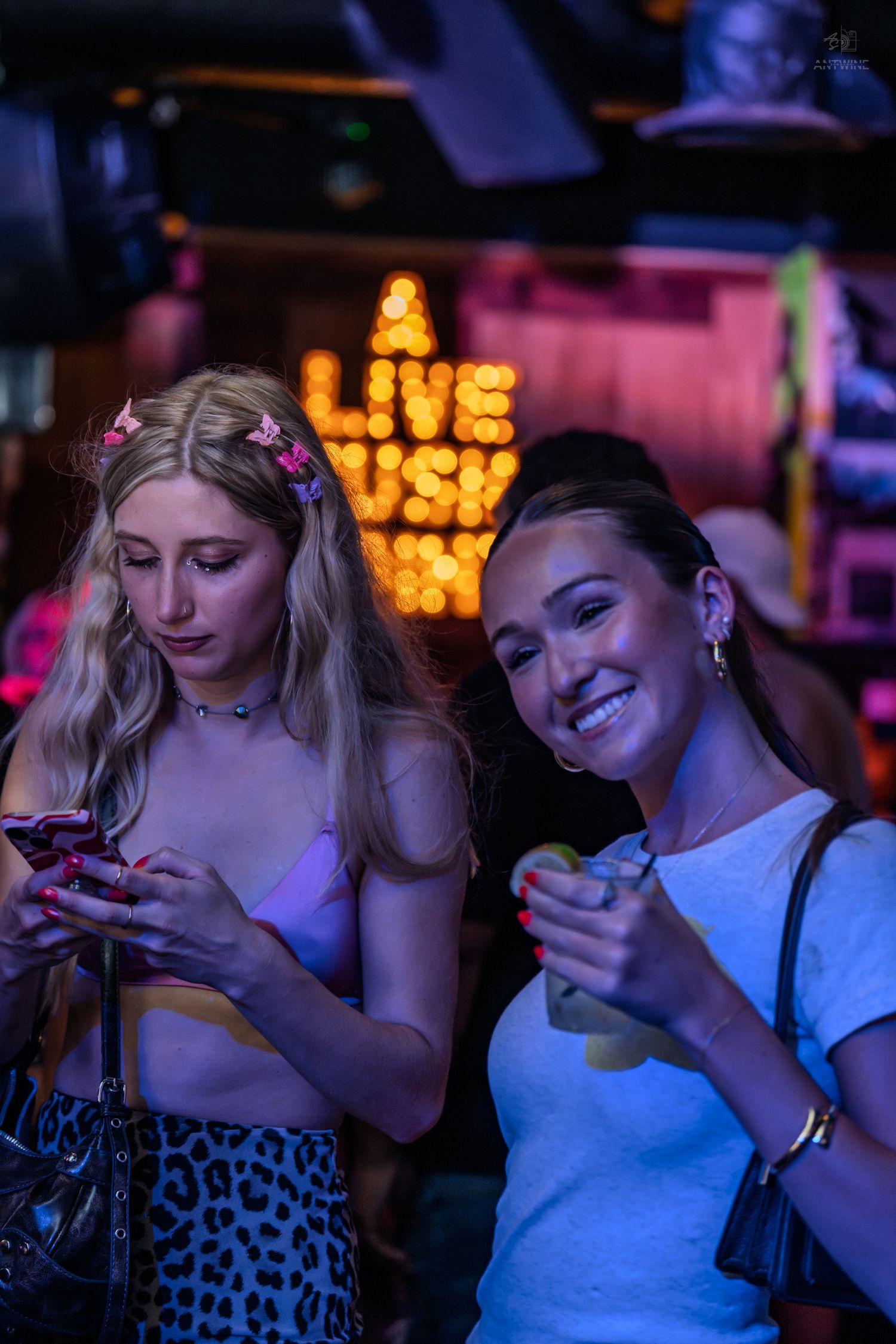 Two young women at a live music bar, one smiling with a drink, the other checking her phone under neon lights.