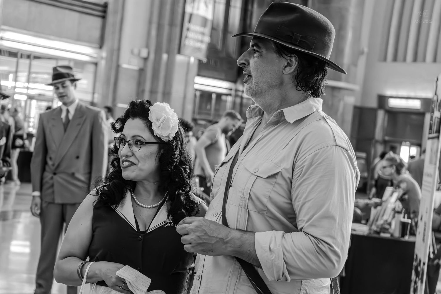 Black and white photo of a couple in vintage 1940s attire at an indoor event, man wearing fedora hat.