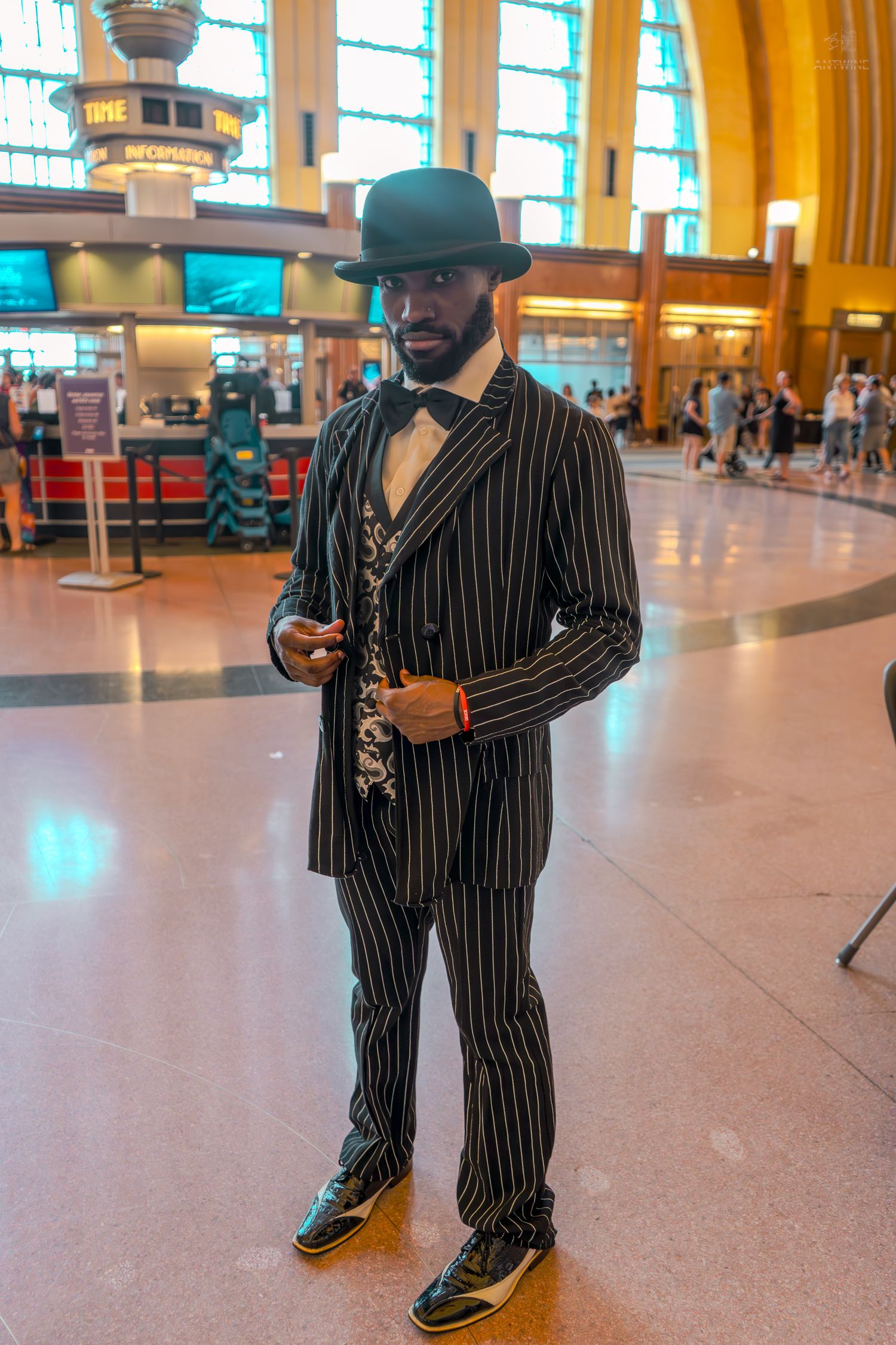 Man in pinstripe suit and bowler hat posing confidently inside a busy train station.