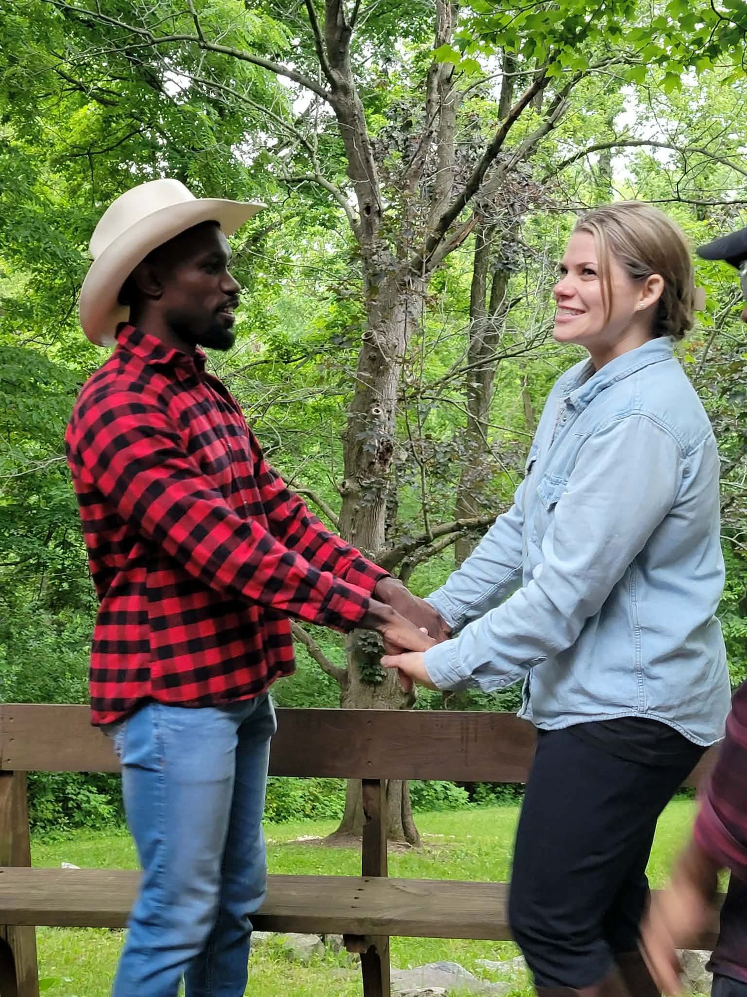 A man in a red plaid shirt and cowboy hat holds hands with a woman in a blue shirt on a wooden deck outdoors.