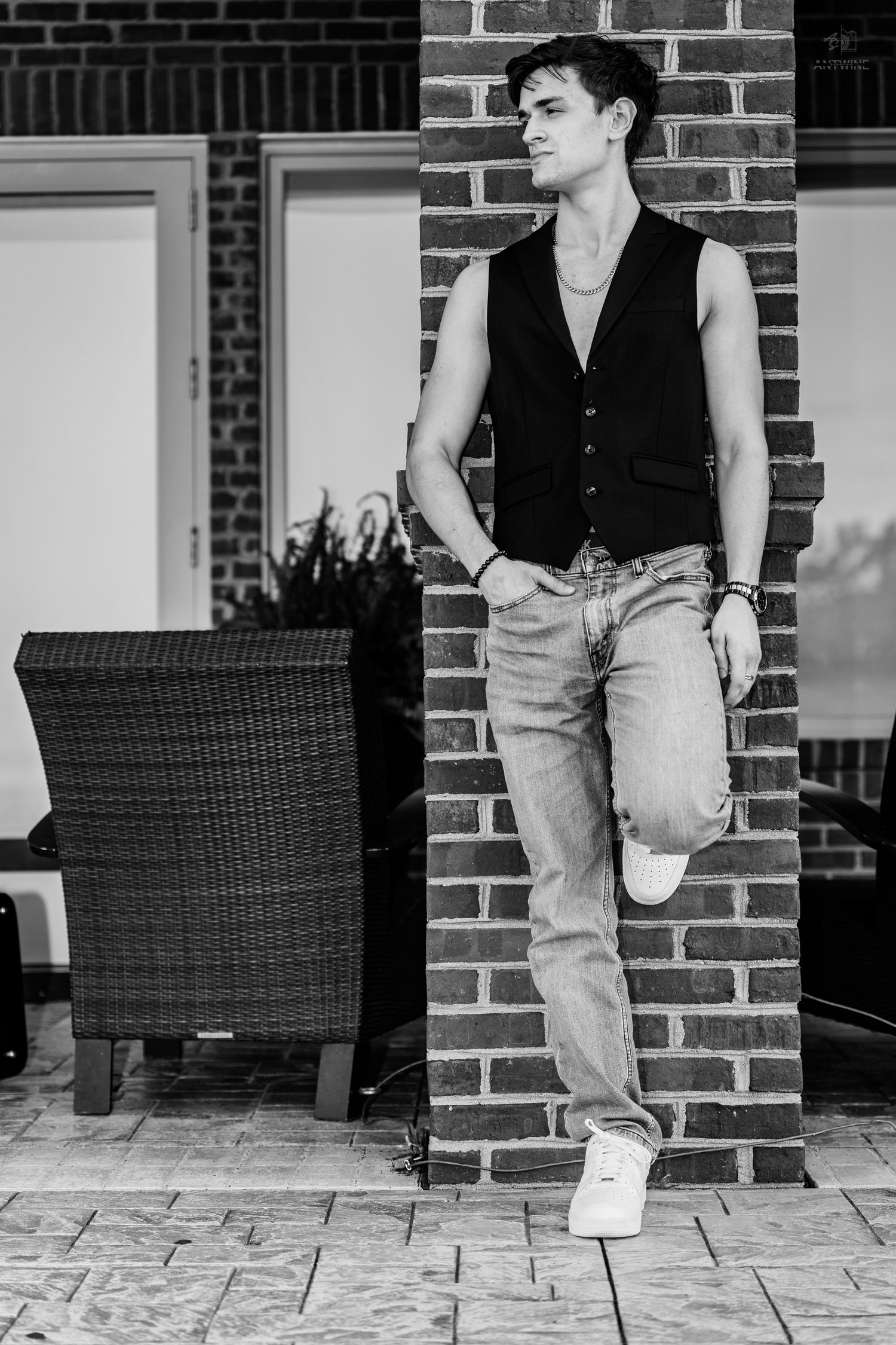 Young man in sleeveless vest and jeans leaning against brick column on patio in black and white photo.
