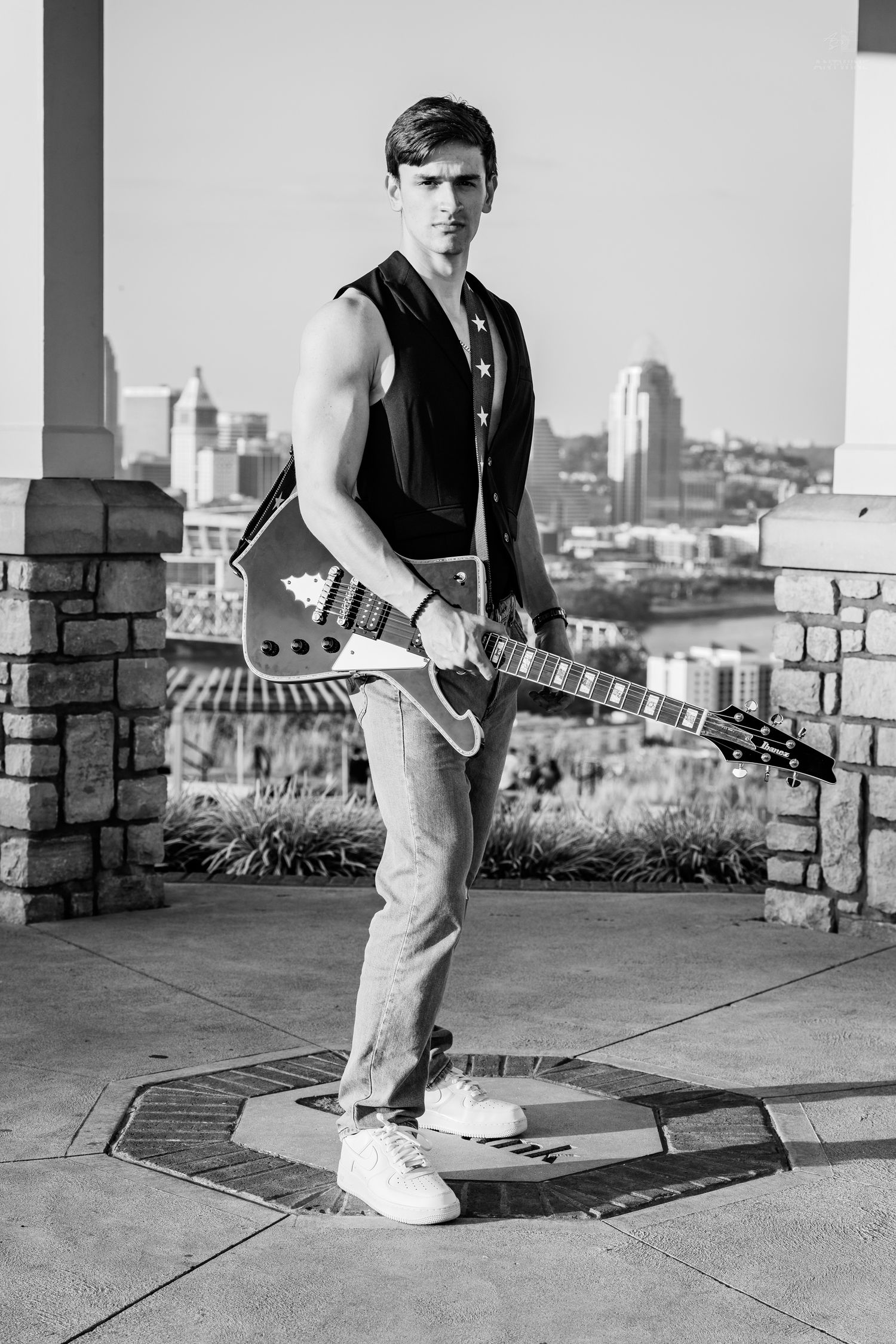 Black and white photo of a muscular young man holding an electric guitar outdoors with a city skyline backdrop.