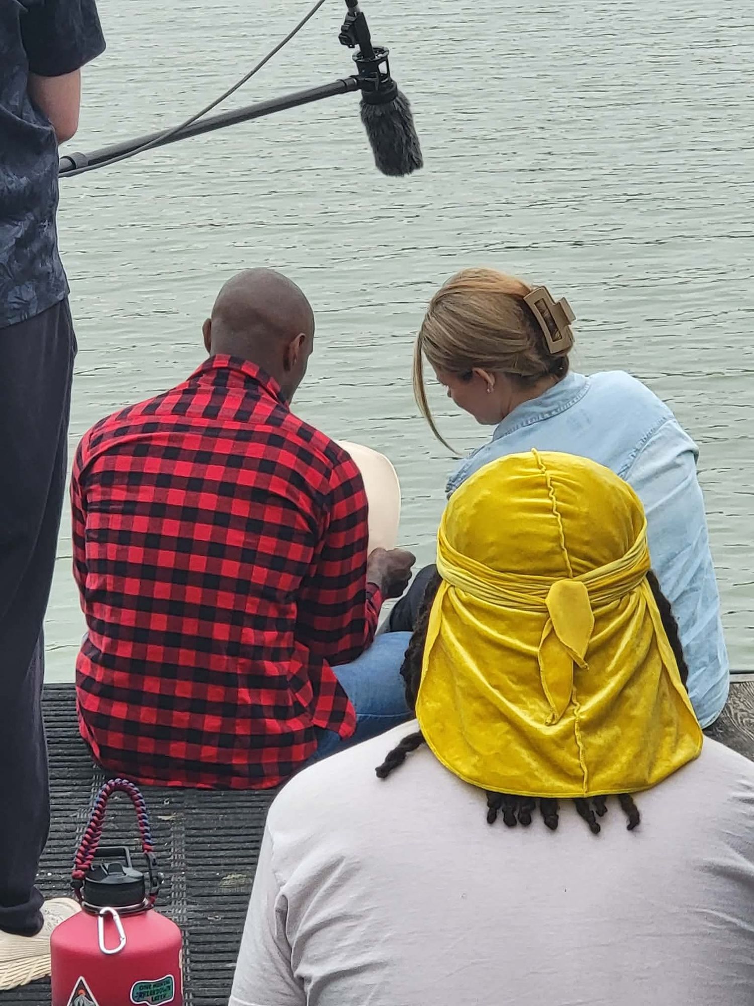 Two people in plaid and yellow hat sit on dock reviewing notes near water with boom mic overhead.