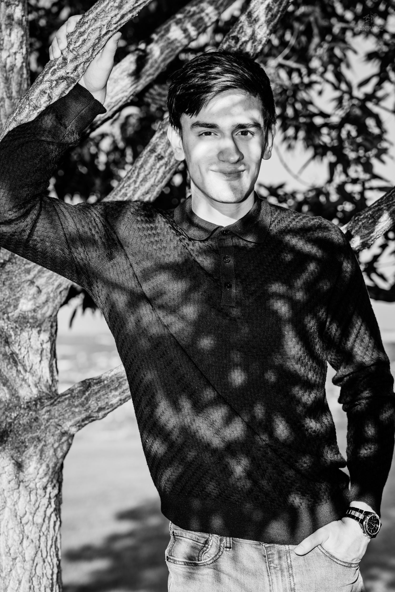 Black and white portrait of a young man leaning against a tree, wearing a sweater and jeans.