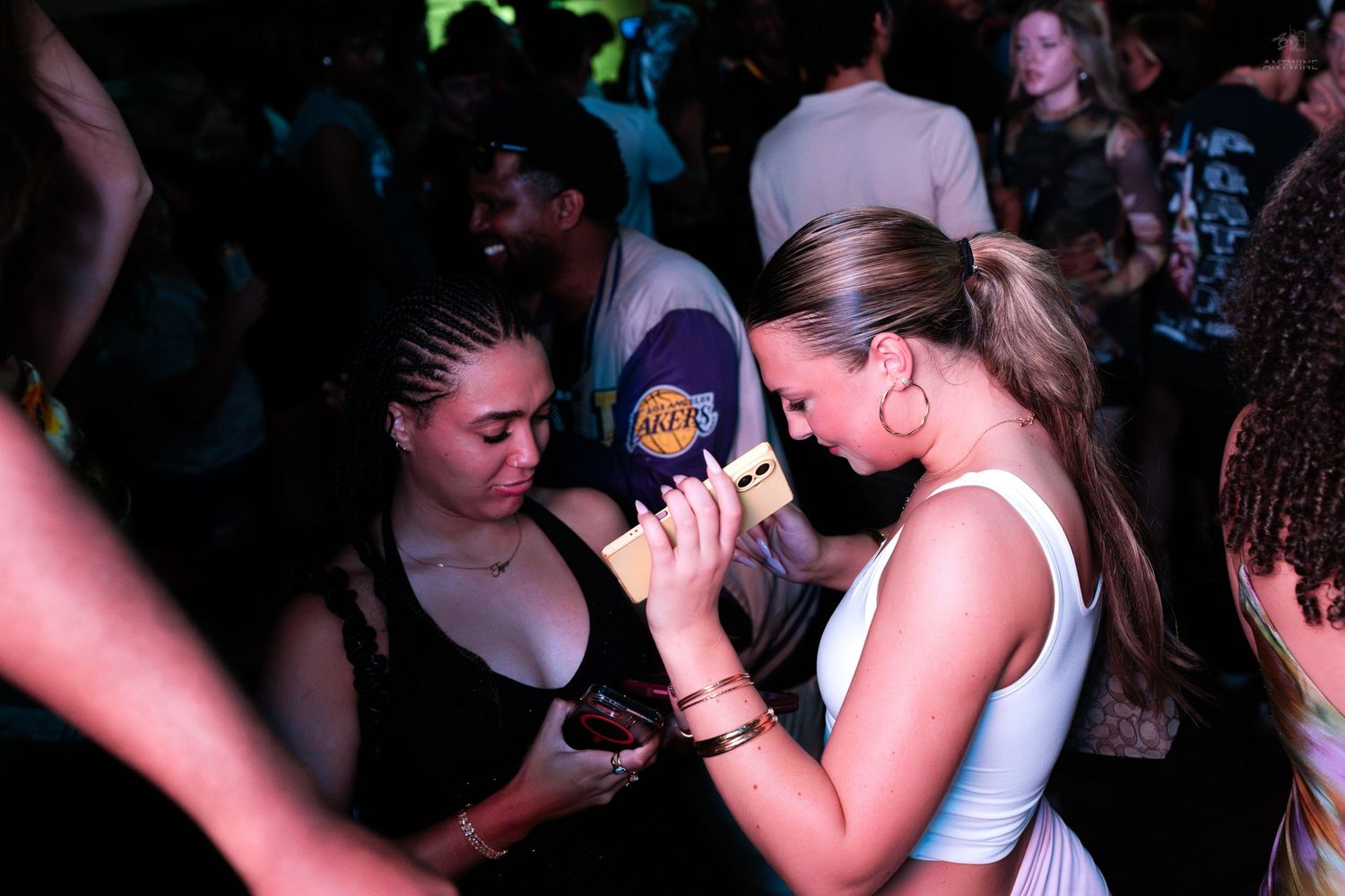 Two women looking at a smartphone together on a busy nightclub dance floor.