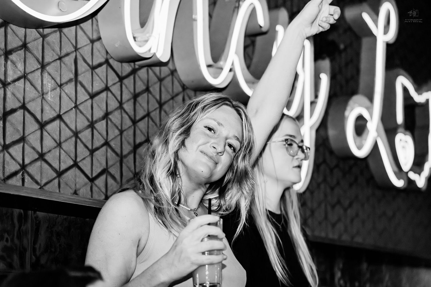 Two women posing at a bar in black and white photo with neon sign backdrop.