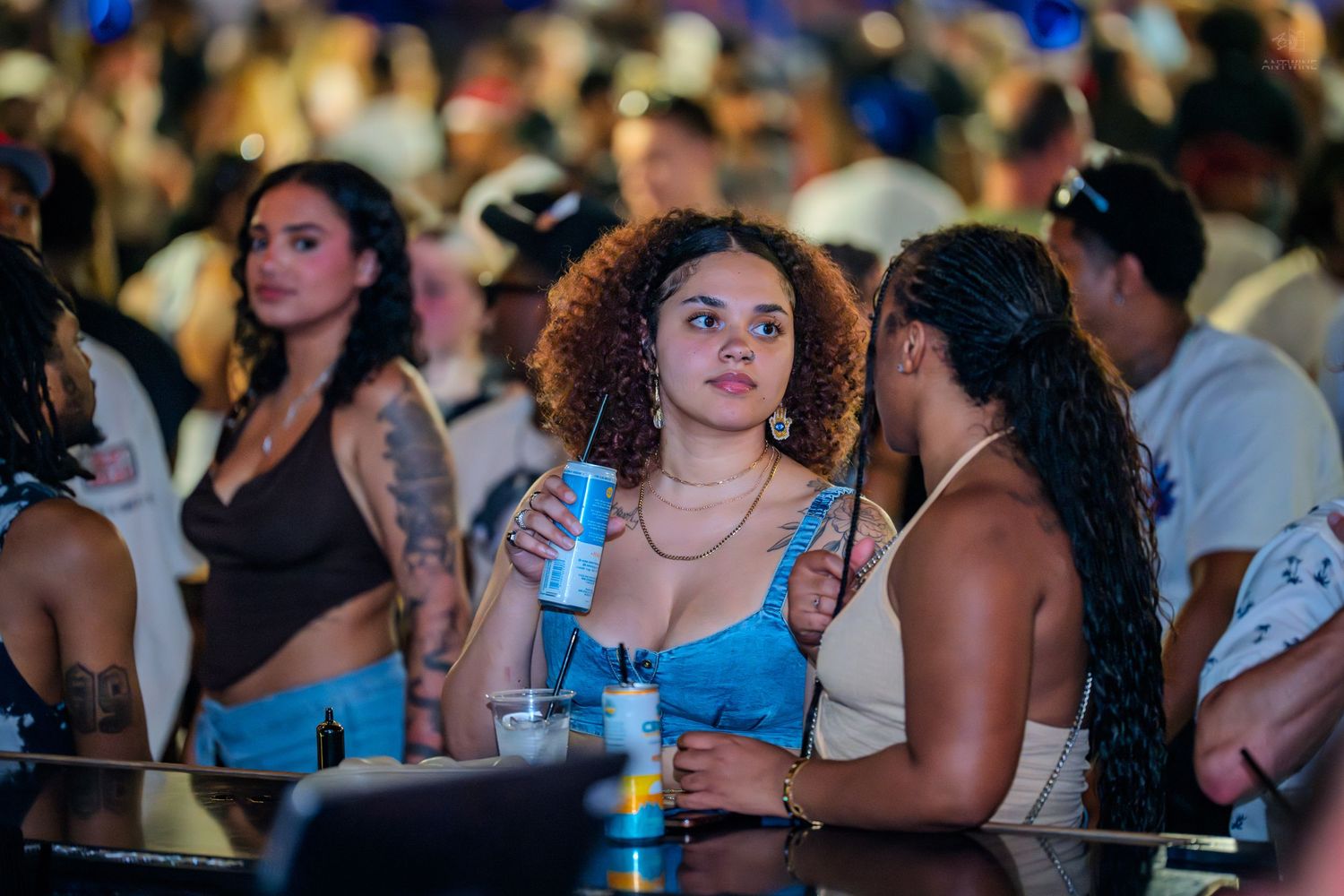Curly-haired woman holds a drink at a lively outdoor nighttime event.
