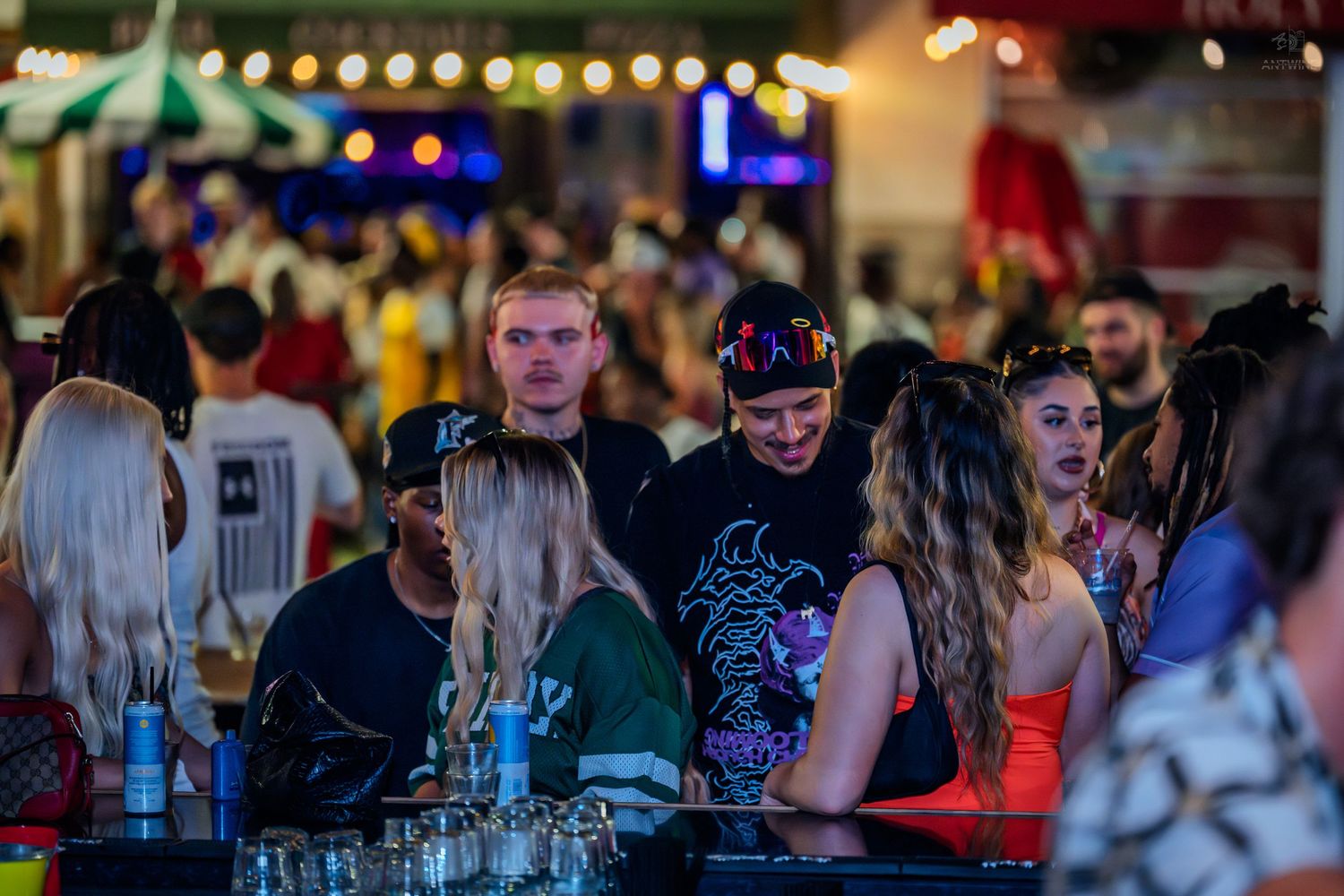 Crowd of young adults socializing at an outdoor nightclub bar event with string lights.