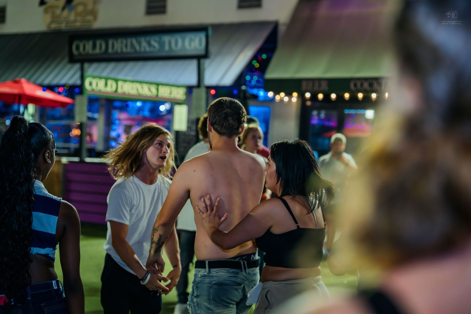 Young adults dancing and socializing outside a Cold Drinks To Go bar at night.