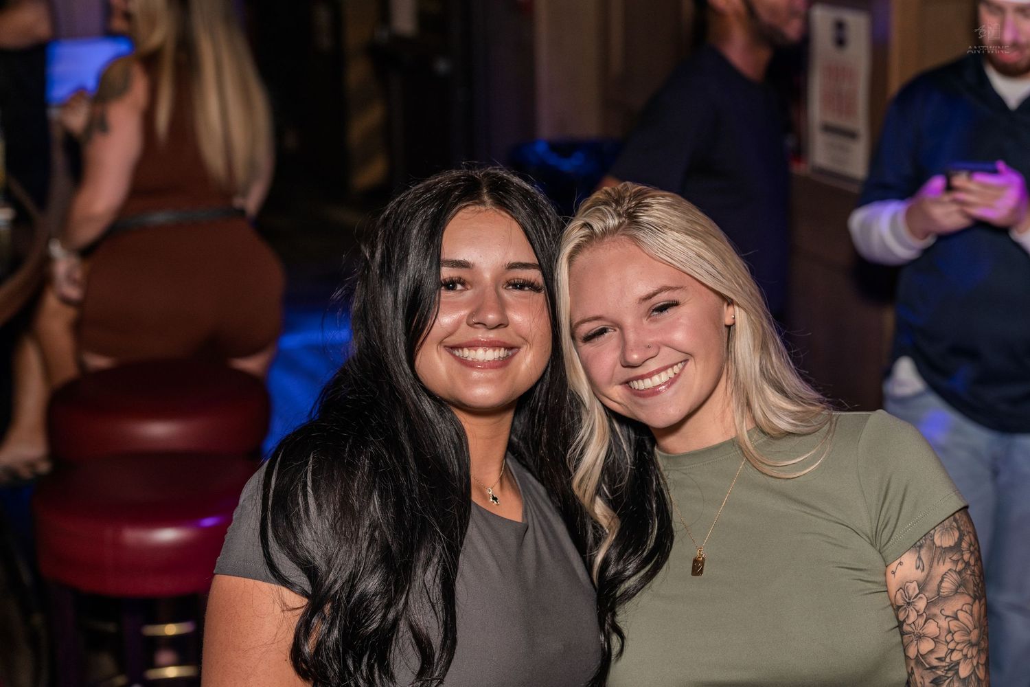 Two smiling women, one brunette and one blonde, posing together at a lively bar event.