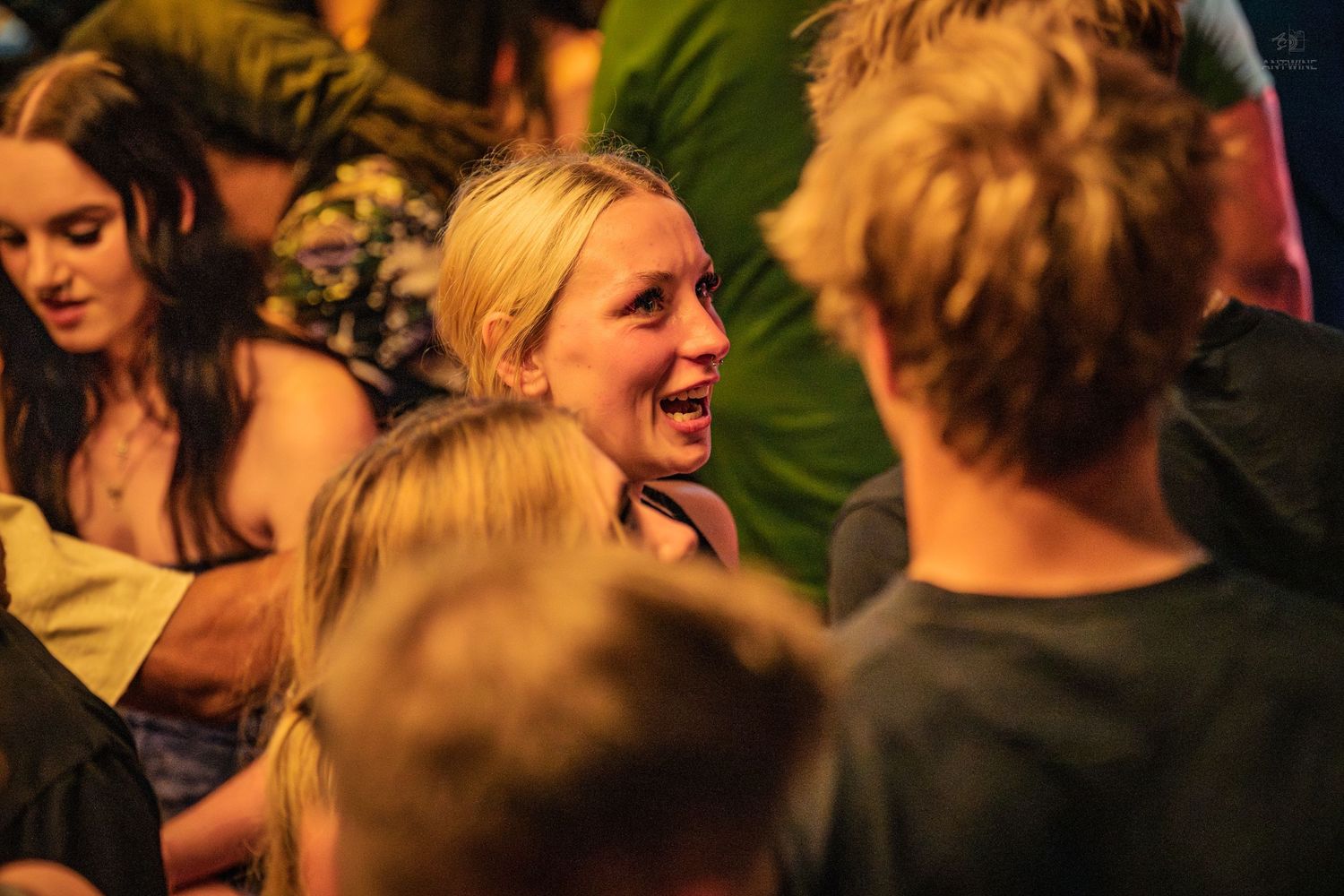 Crowded indoor social event with people mingling, featuring a tall man in a black t-shirt at center.