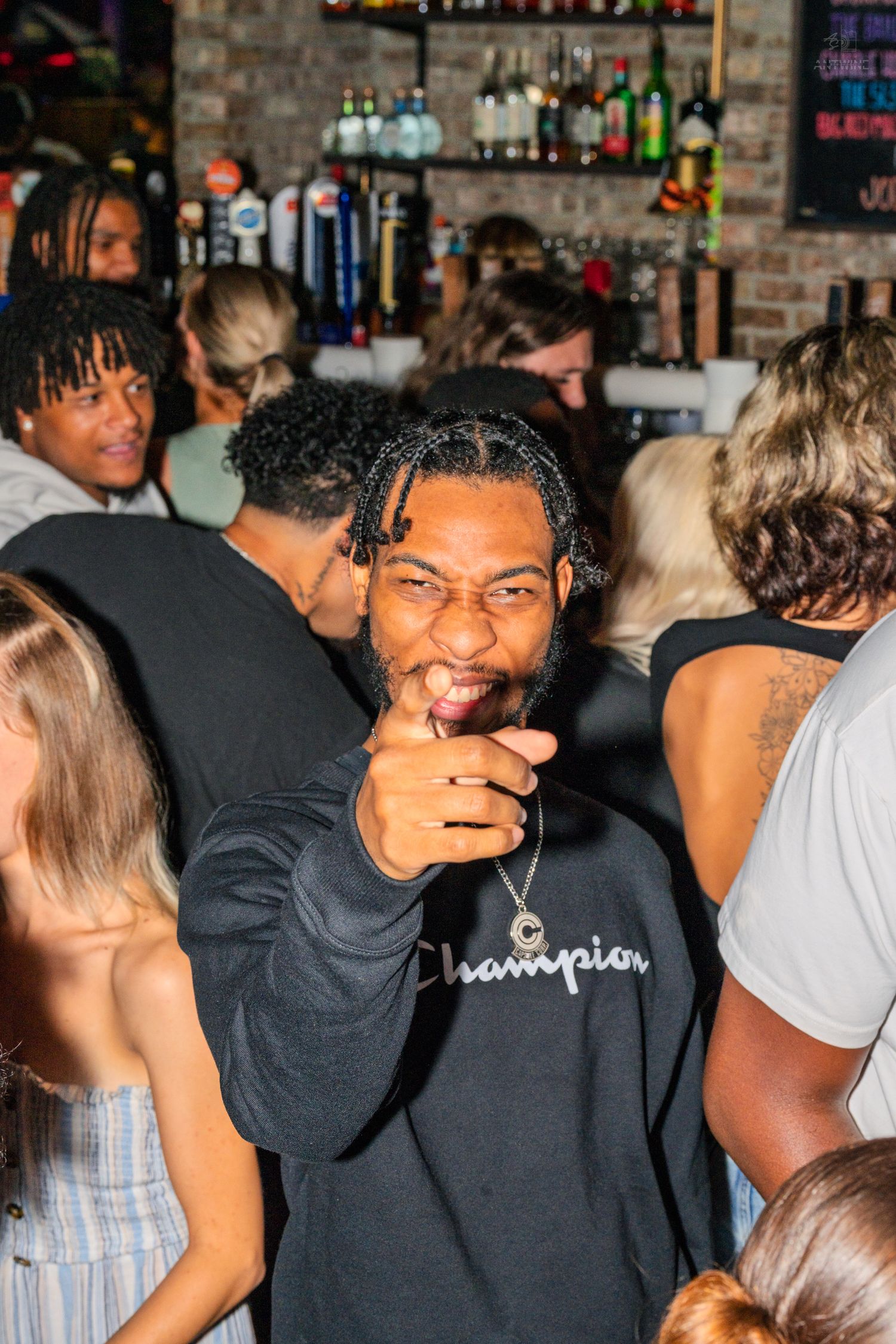 Man in Champion sweatshirt smiling and pointing at camera in a crowded bar.