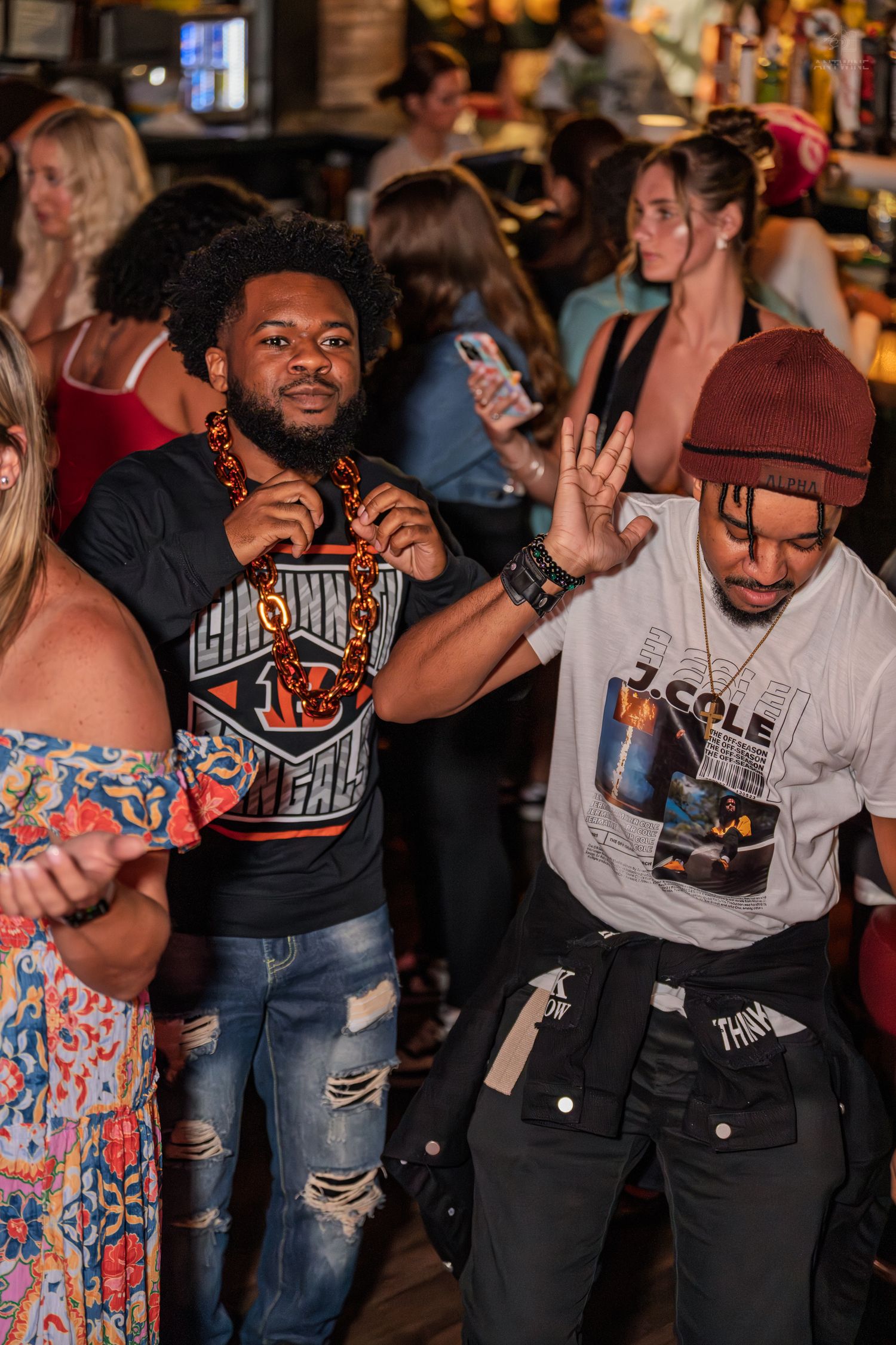 Two men dancing at a vibrant bar party, one wearing a J. Cole graphic tee.