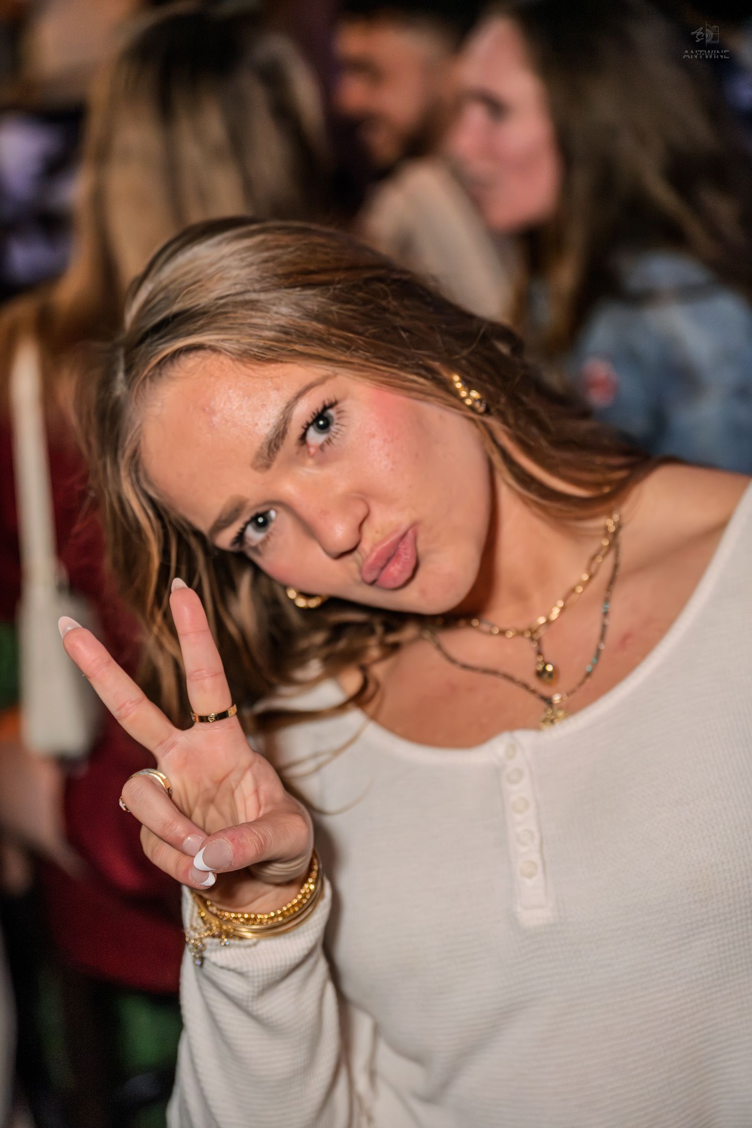 A young woman flashes a peace sign and playful expression at a crowded nightclub.