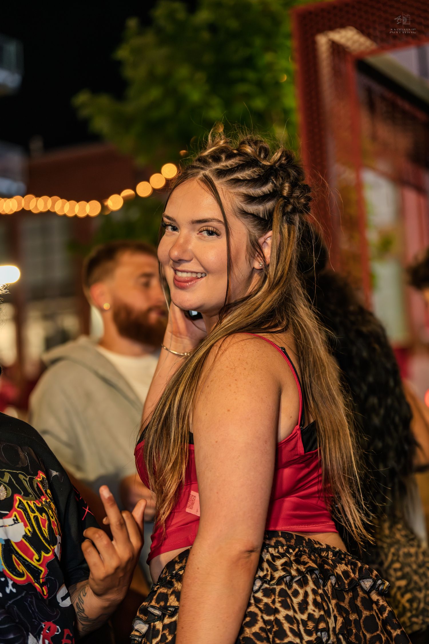 Young woman with braided updo hairstyle smiling at night outdoor event wearing red top and leopard print skirt.