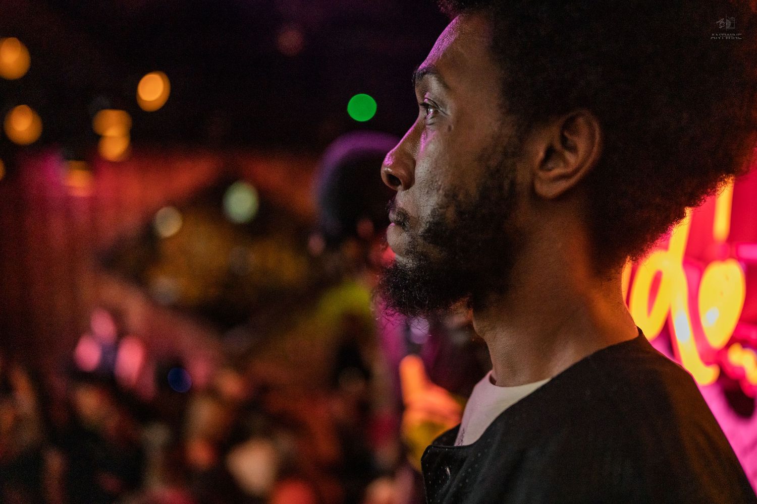 Side profile of a bearded Black man with an afro at a lively nightclub with pink and green neon lights.