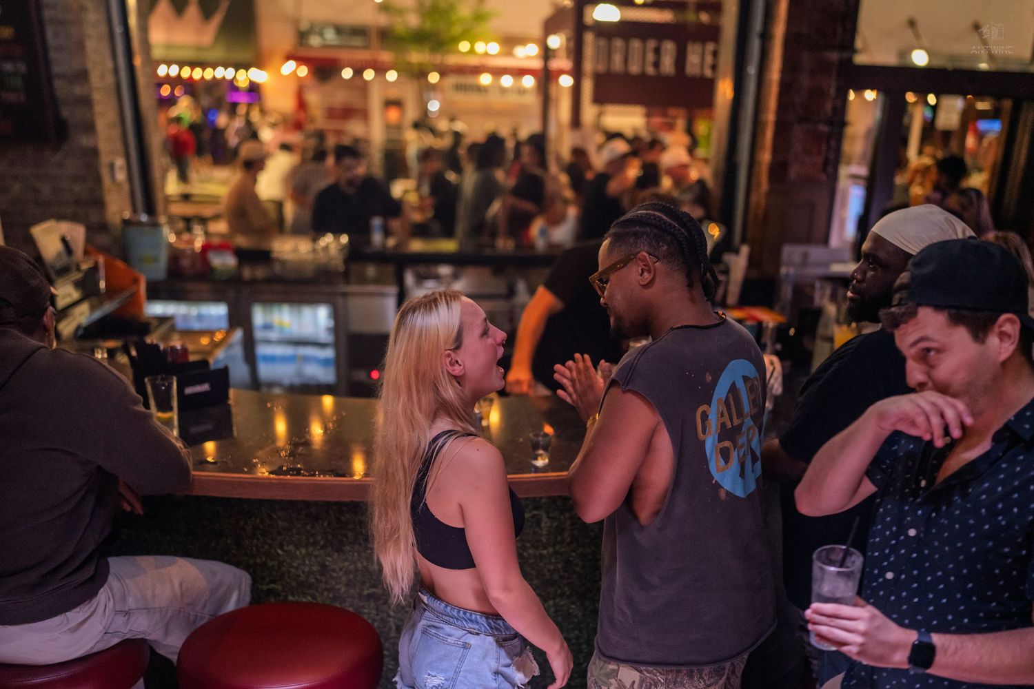 Blonde woman and man share an intimate conversation at a vibrant nightclub bar with glowing string lights overhead.