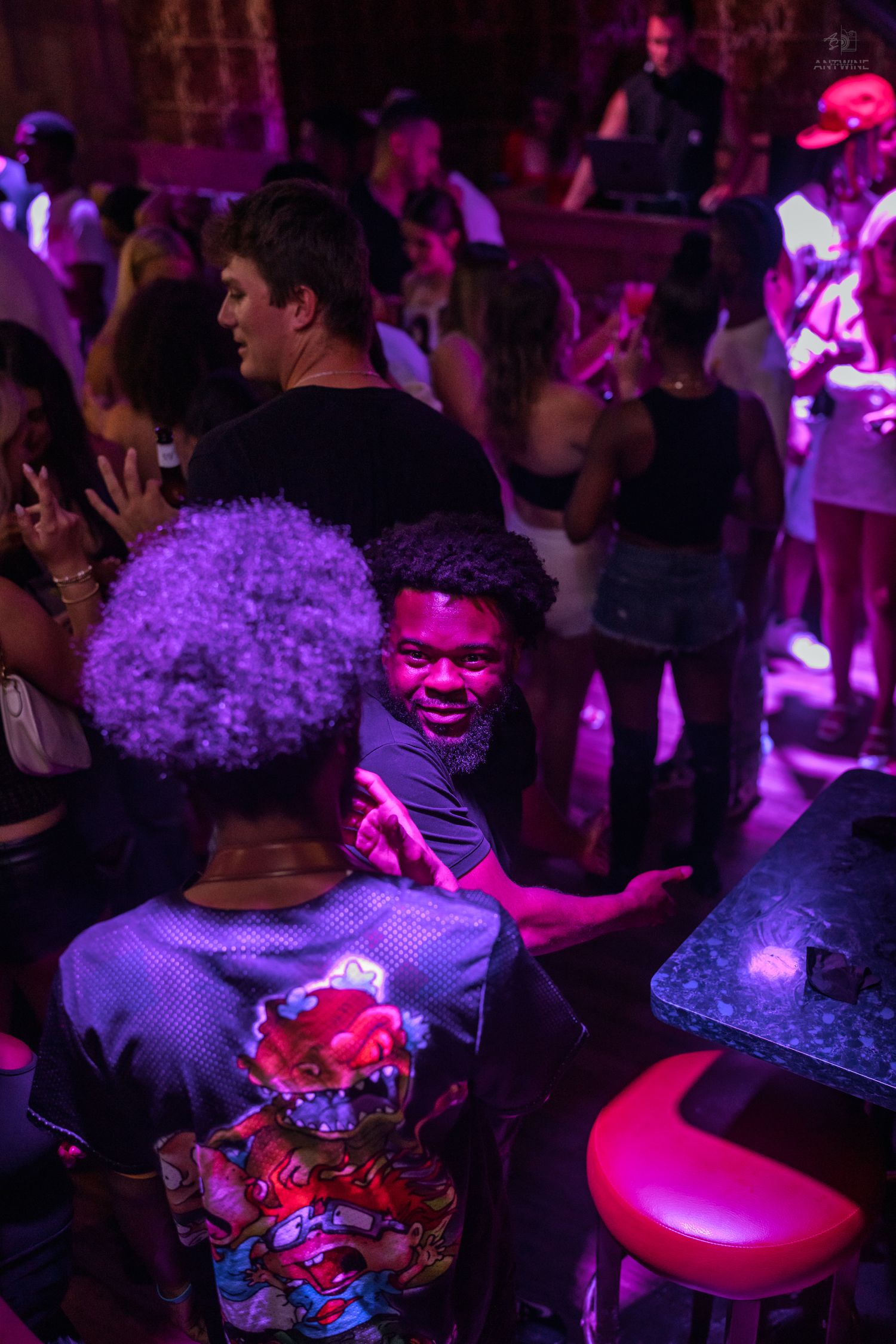 Man smiling under pink and purple neon lights at a vibrant crowded nightclub party.