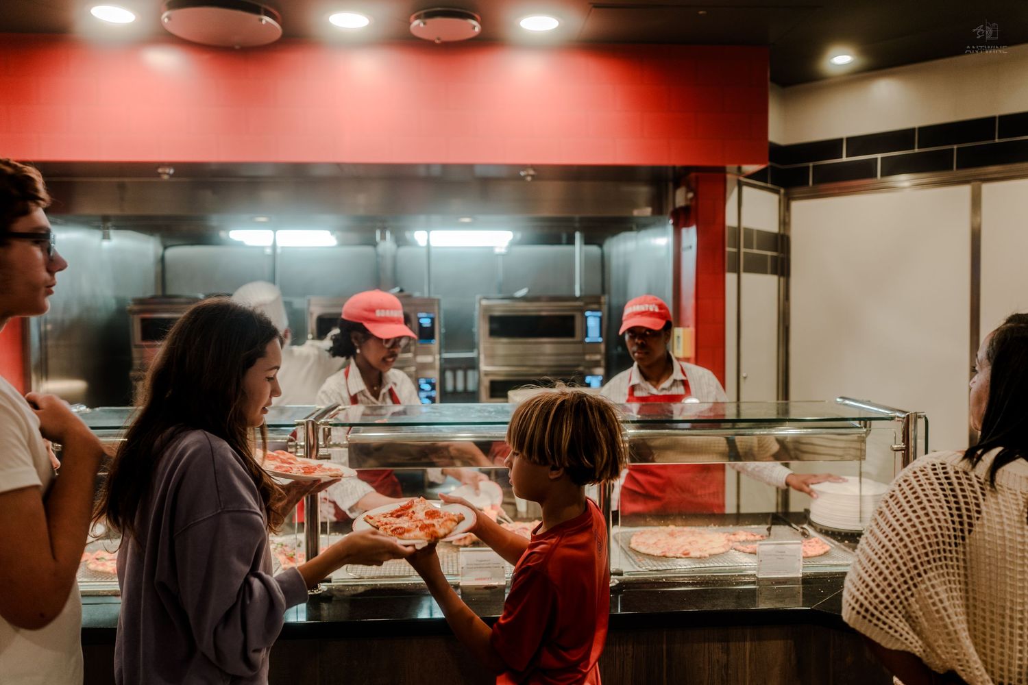 Customers ordering pizza at a fast food restaurant counter with staff in red uniforms serving slices.