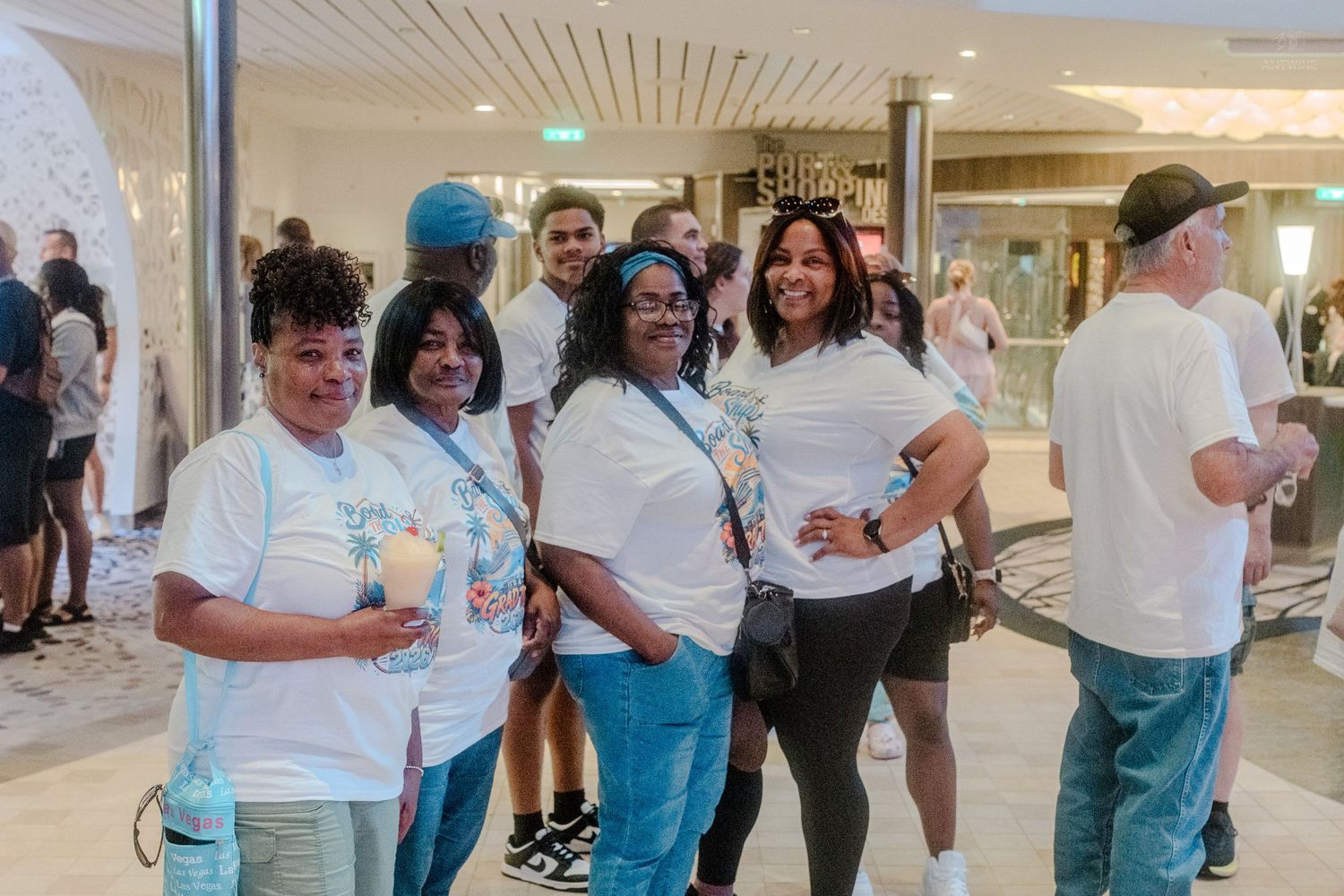 Group of four women in matching white cruise t-shirts posing together in a ship interior.