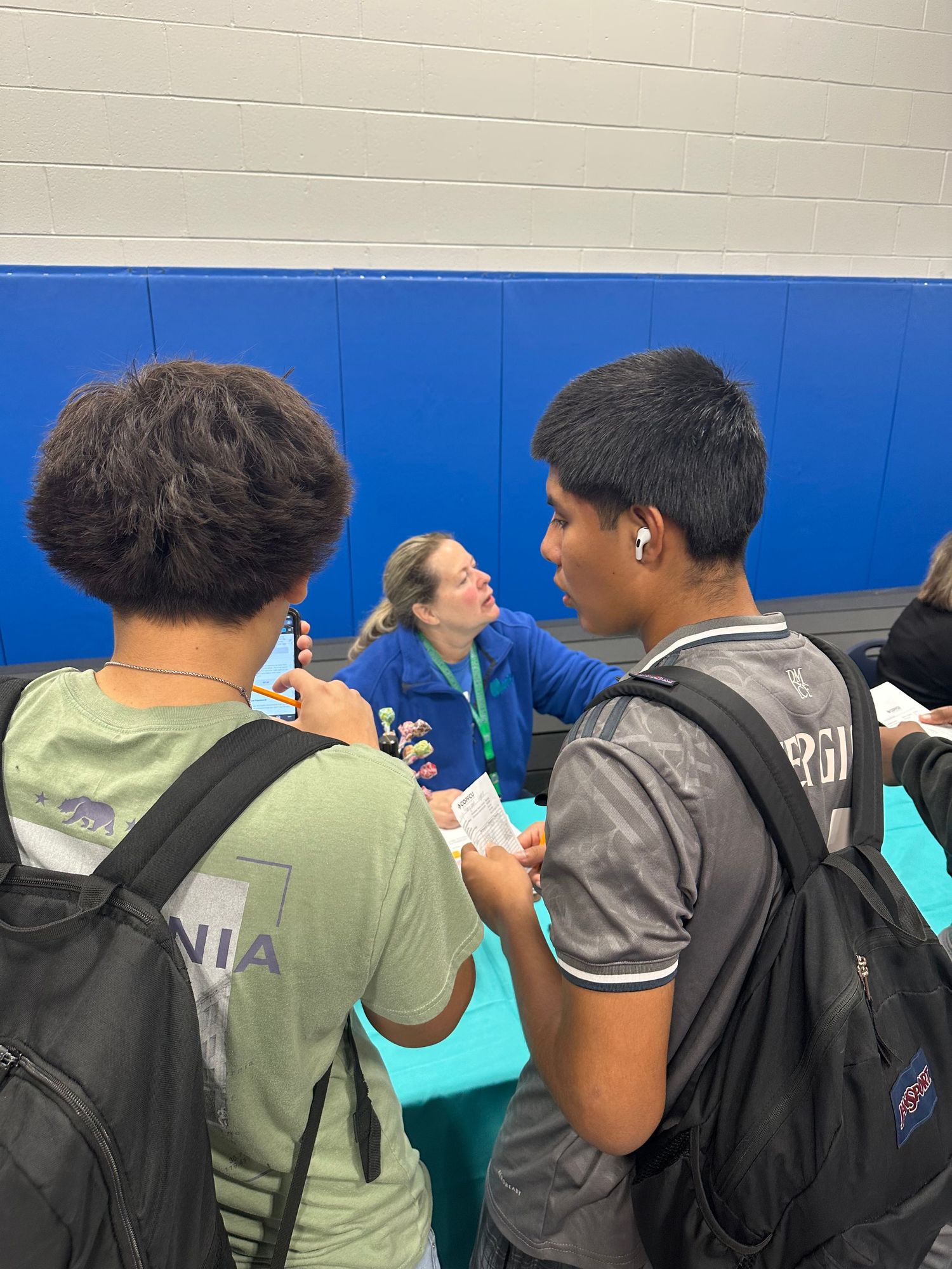 Two students with backpacks speak with a woman at a blue-walled school gymnasium information table.