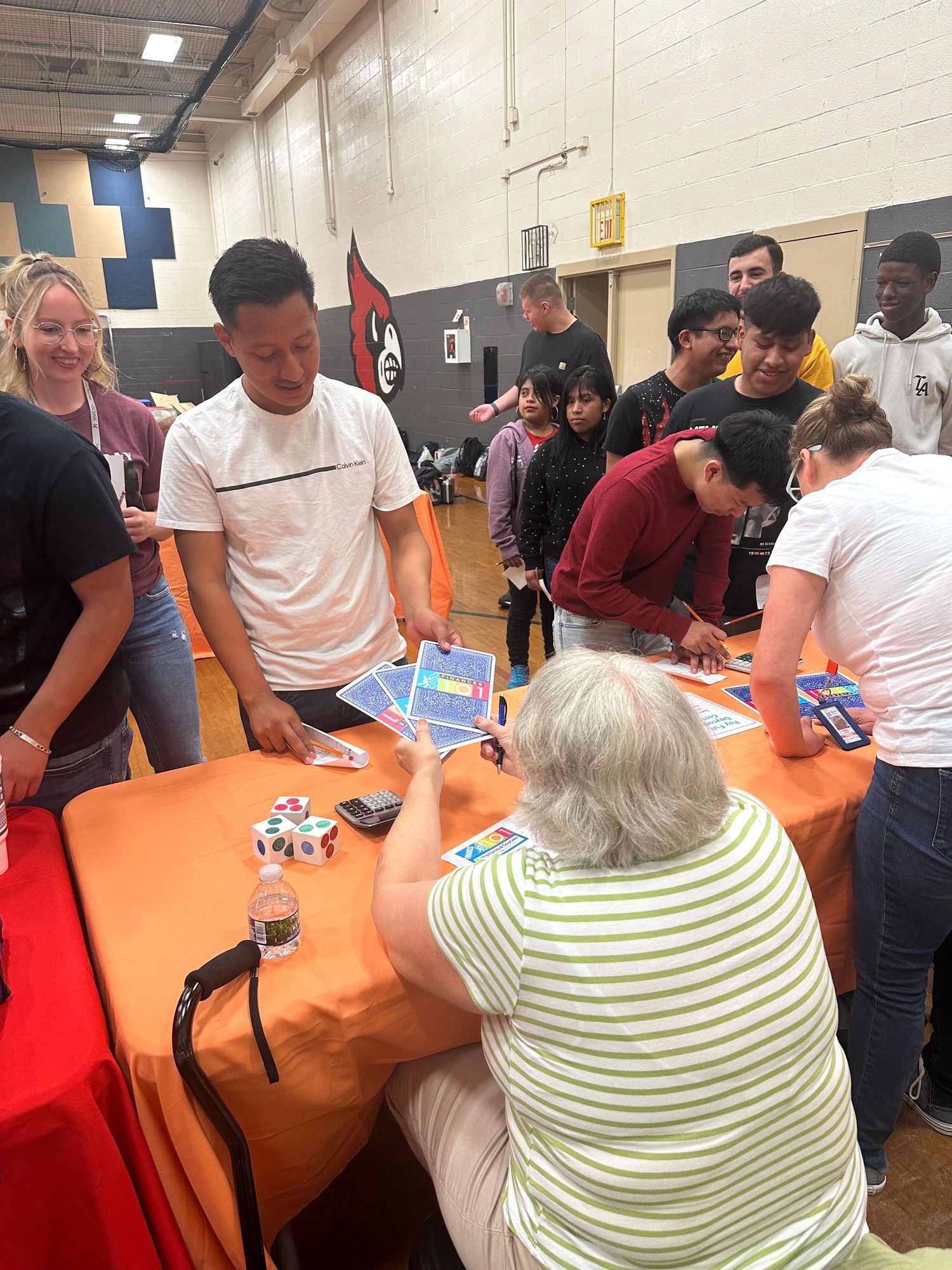 Students and adults gather around an orange table at a school gym event, reviewing colorful papers and materials.