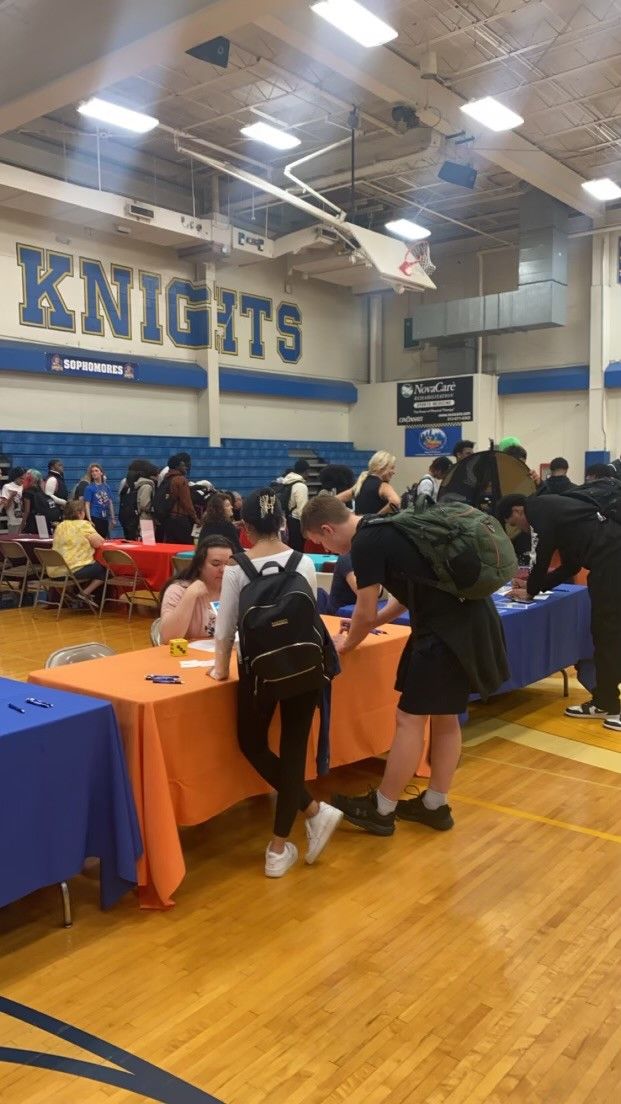 Students gather at colorful tables in a school gymnasium decorated with a Knights banner during a college or career fair.