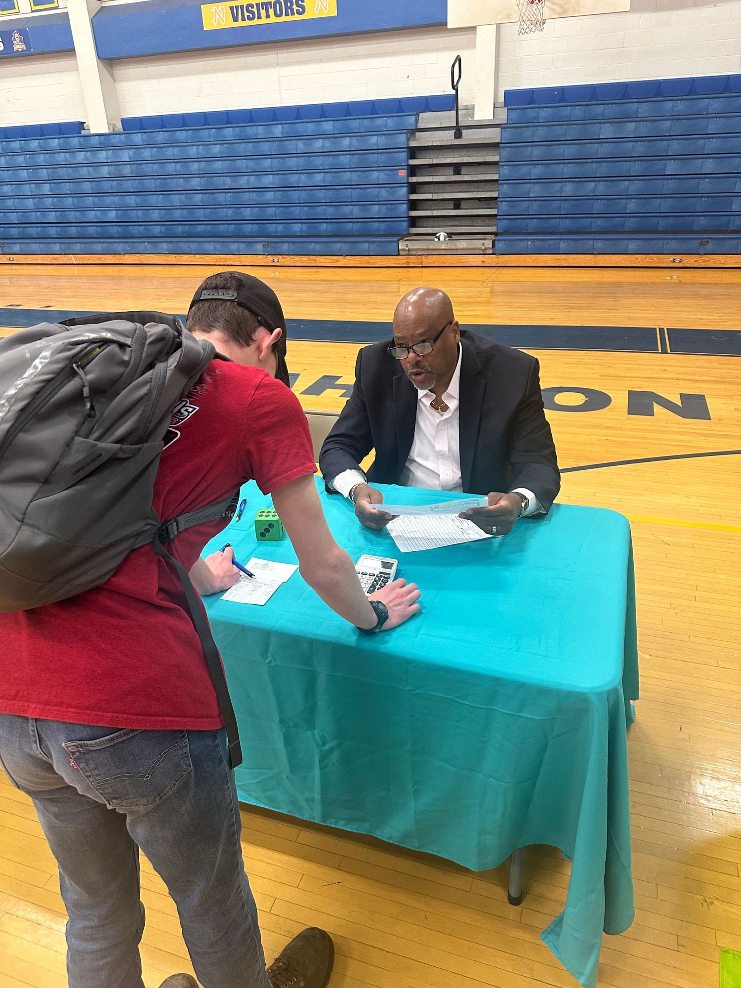 Man in suit sits at teal table in gymnasium speaking with a student carrying a backpack at a career fair.
