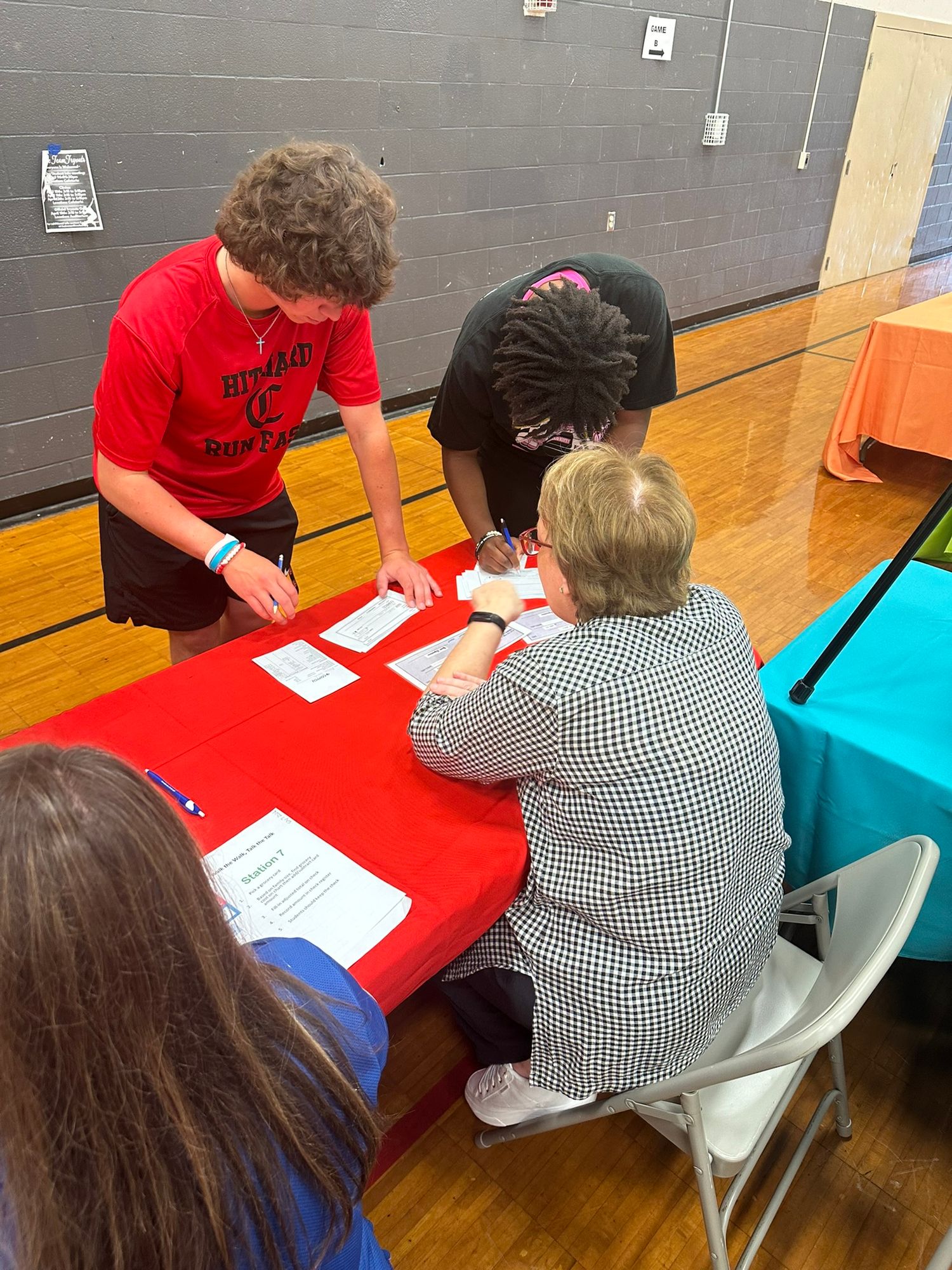 Two students and an adult woman review and sign documents at a red table inside a school gymnasium during an event.