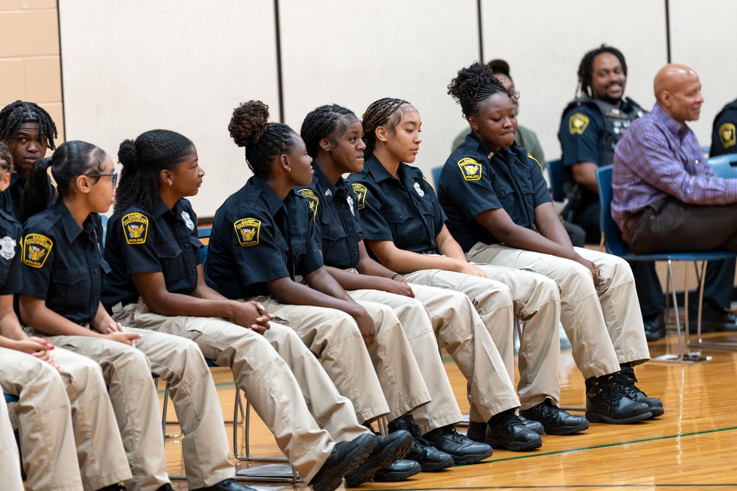 Young women in matching black uniforms and khaki pants sit in a row during a formal ceremony in a gymnasium.