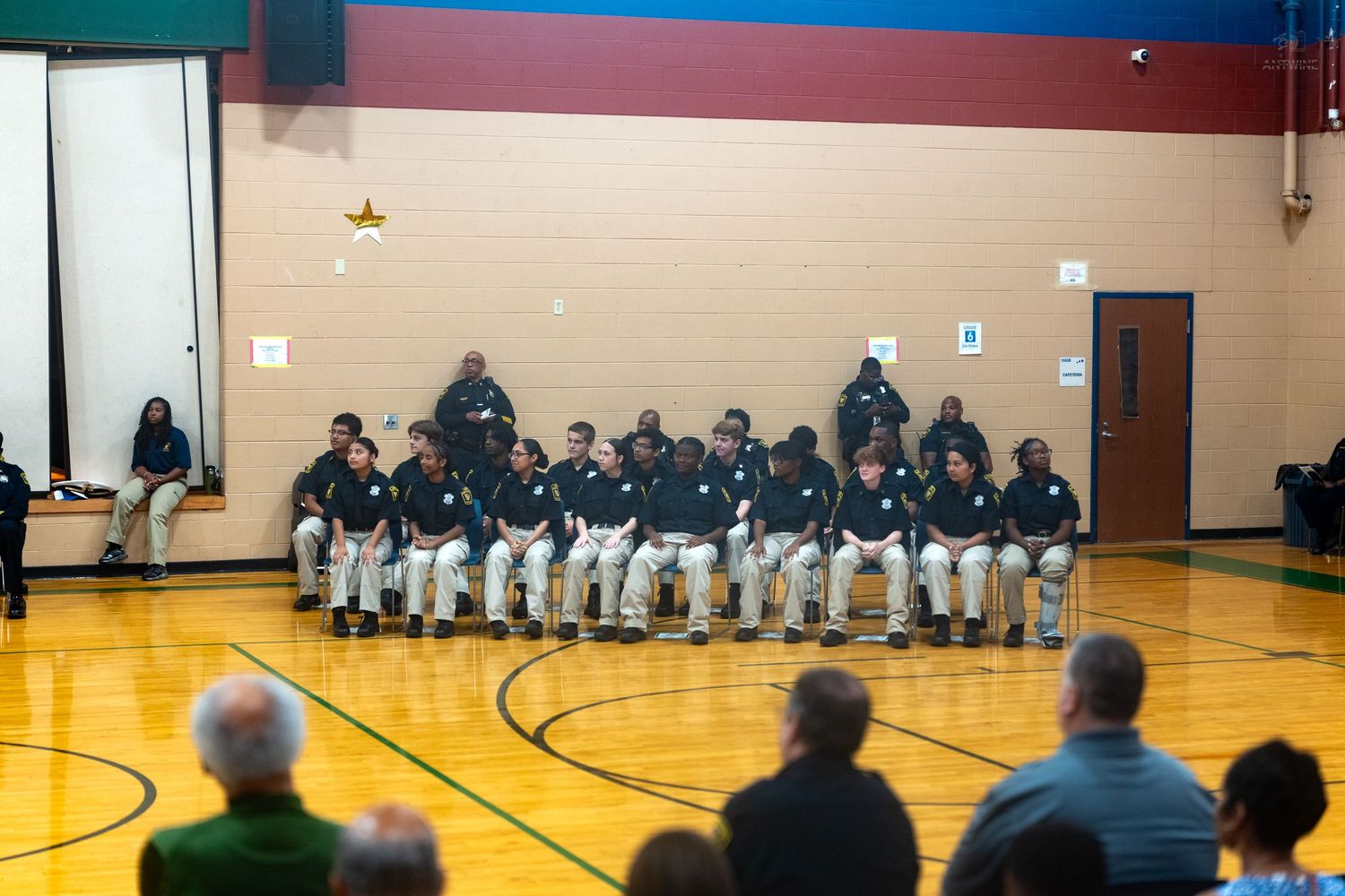 Group of men in matching uniforms seated in chairs on a gymnasium basketball court during a formal indoor event.