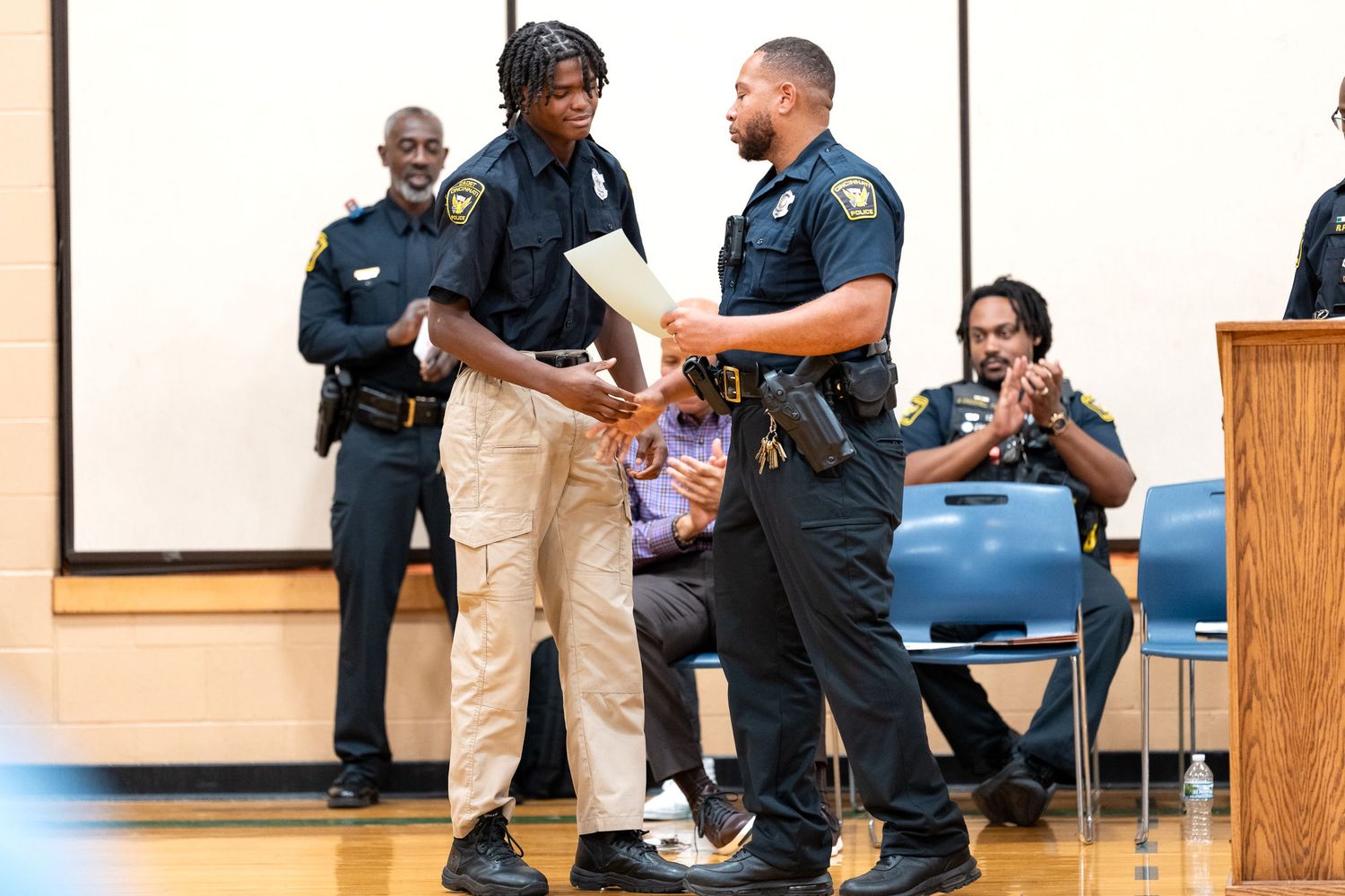 Police officers shake hands with a young man during an award ceremony in a school gymnasium.
