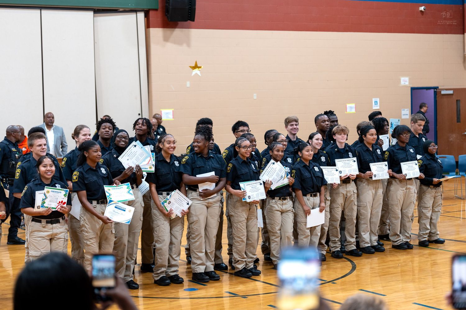 Students in tan uniforms holding certificates at an indoor awards ceremony in a school gymnasium.