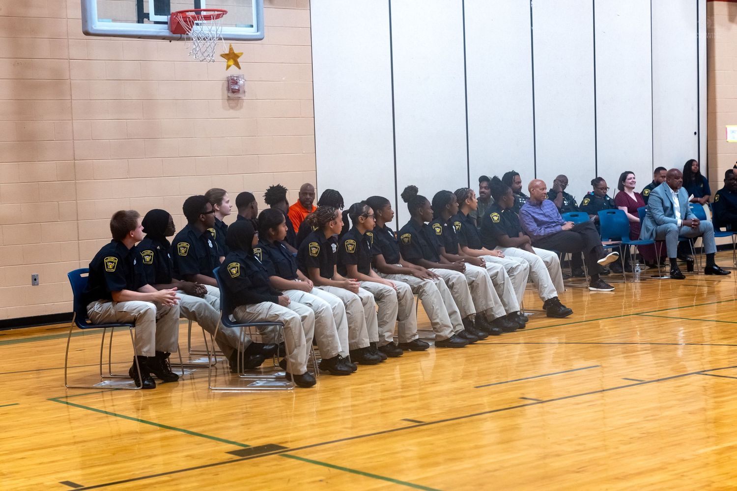 Students in matching uniforms sit in rows on a gym floor during a school assembly or ceremony.