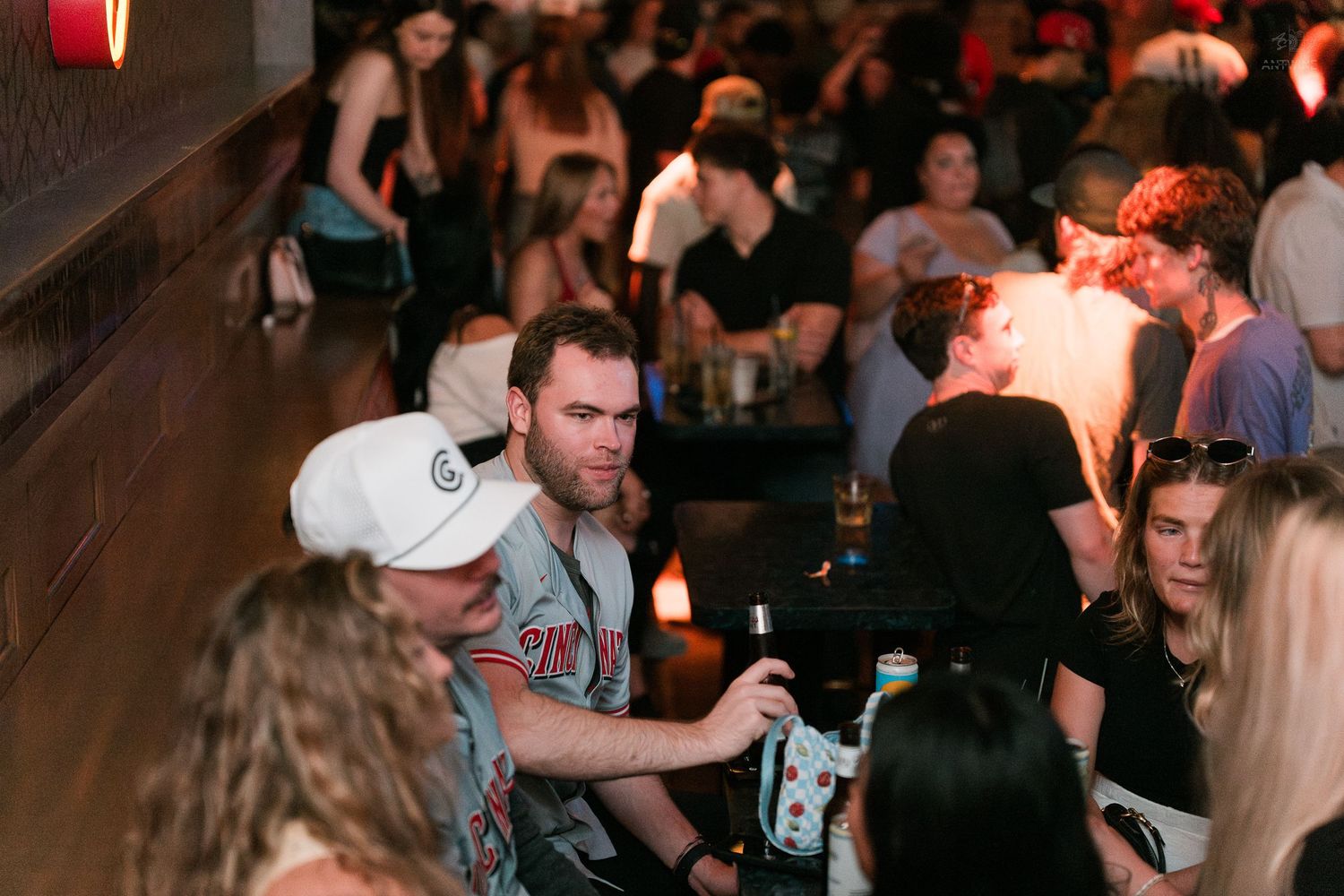 Young man in white cap enjoys drinks with friends at a dimly lit bar counter.