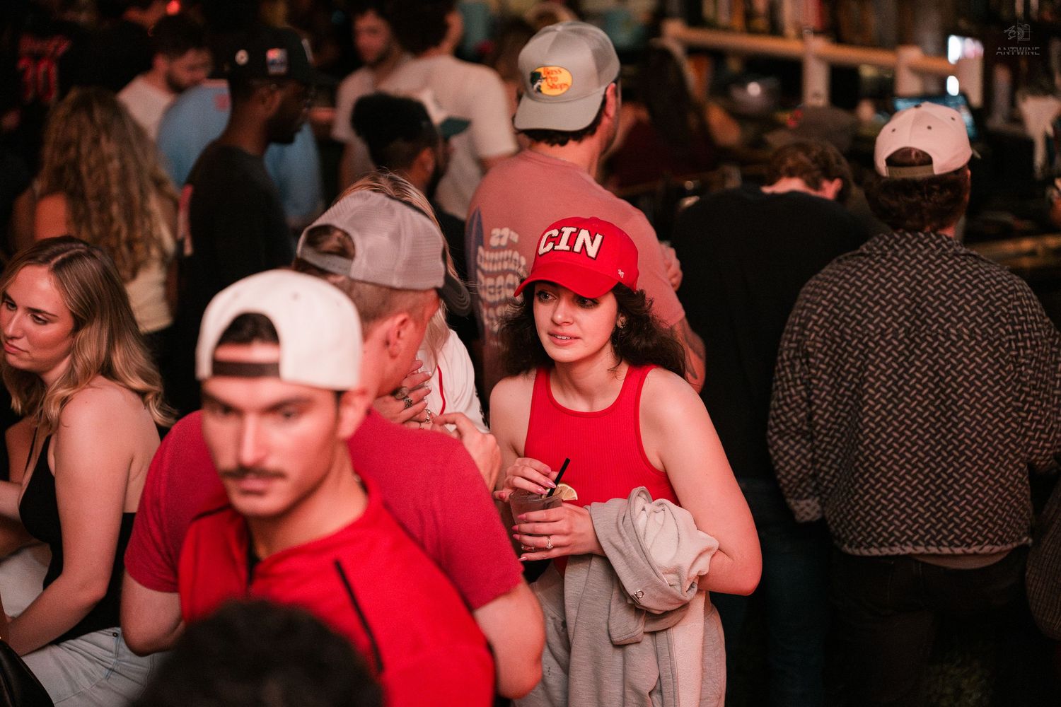 Crowded bar scene with people socializing, woman in red tank top holding drink stands out in busy nightlife venue.