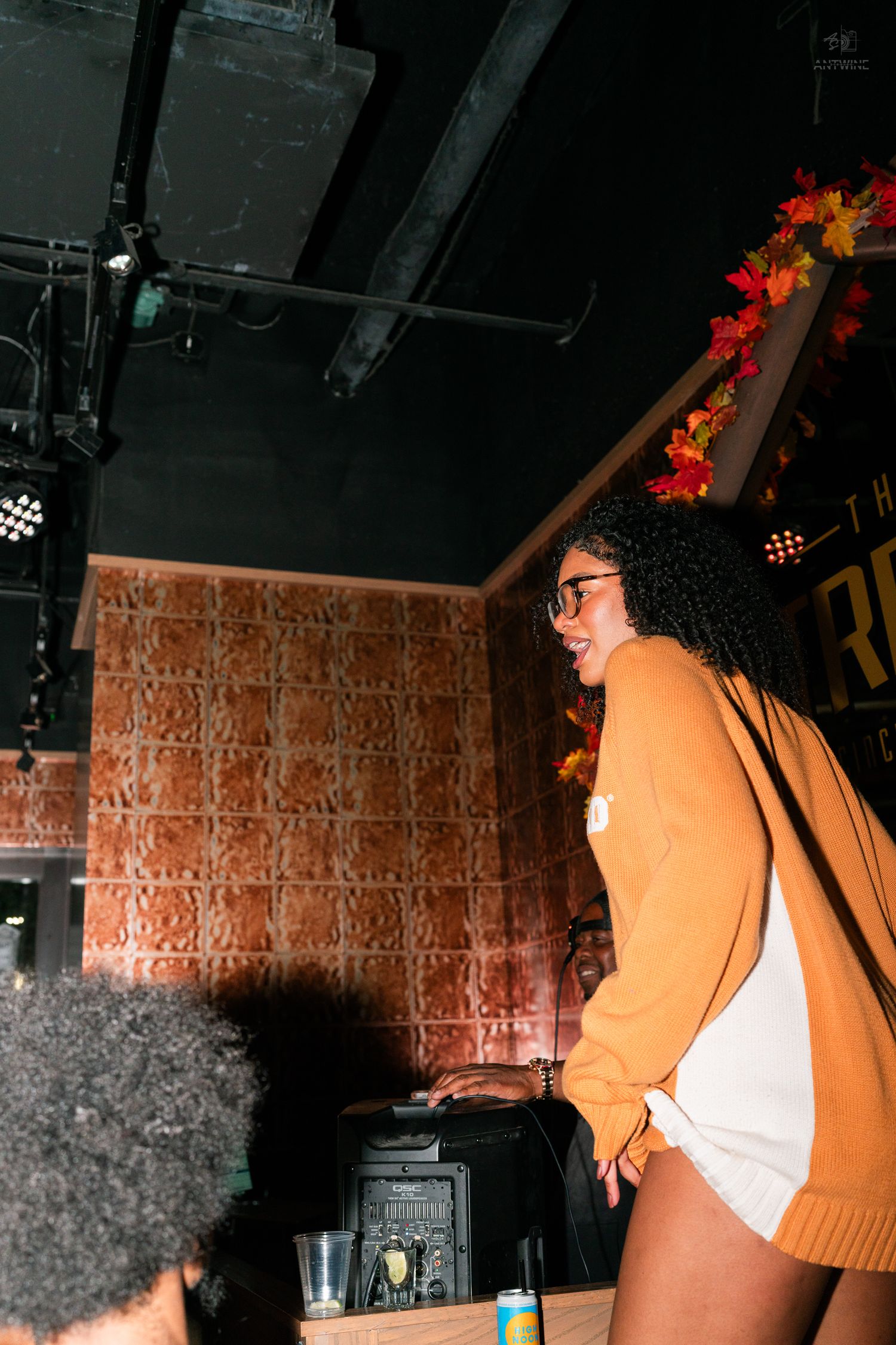 Smiling woman in orange sweater standing at a bar decorated with fall foliage.