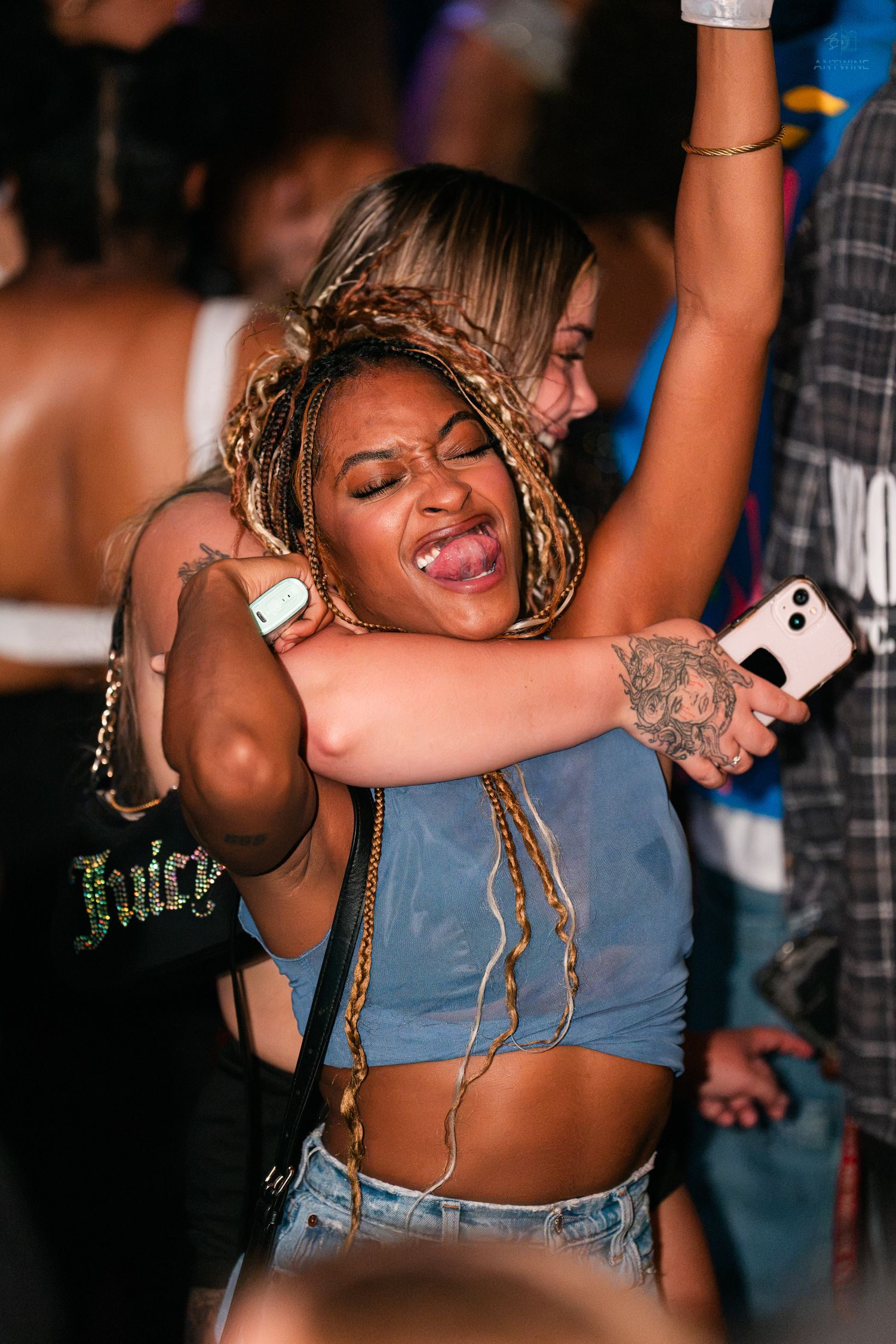 Woman with colorful braids dancing joyfully at a crowded nightclub party, holding a phone.