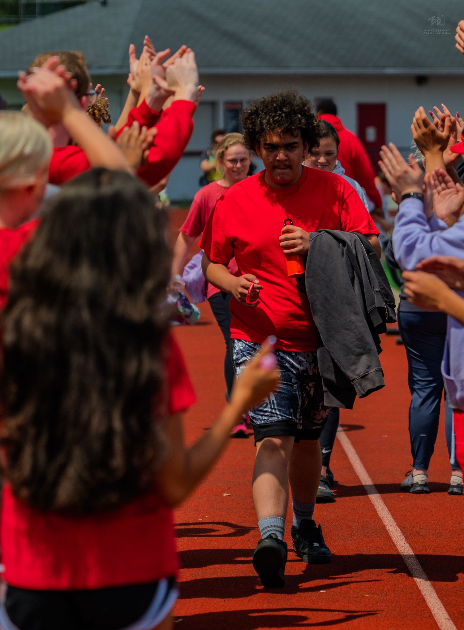 Student runs through cheering crowd on red track during school sports event, surrounded by peers in red shirts.
